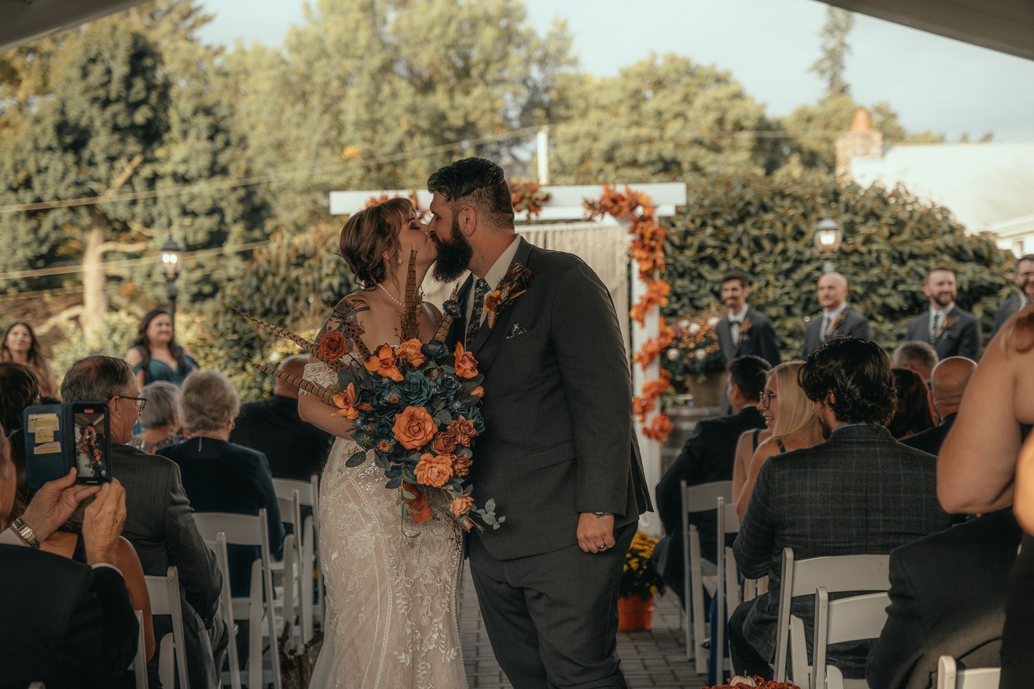Newlyweds share first kiss at outdoor wedding ceremony with orange floral arch and seated guests.