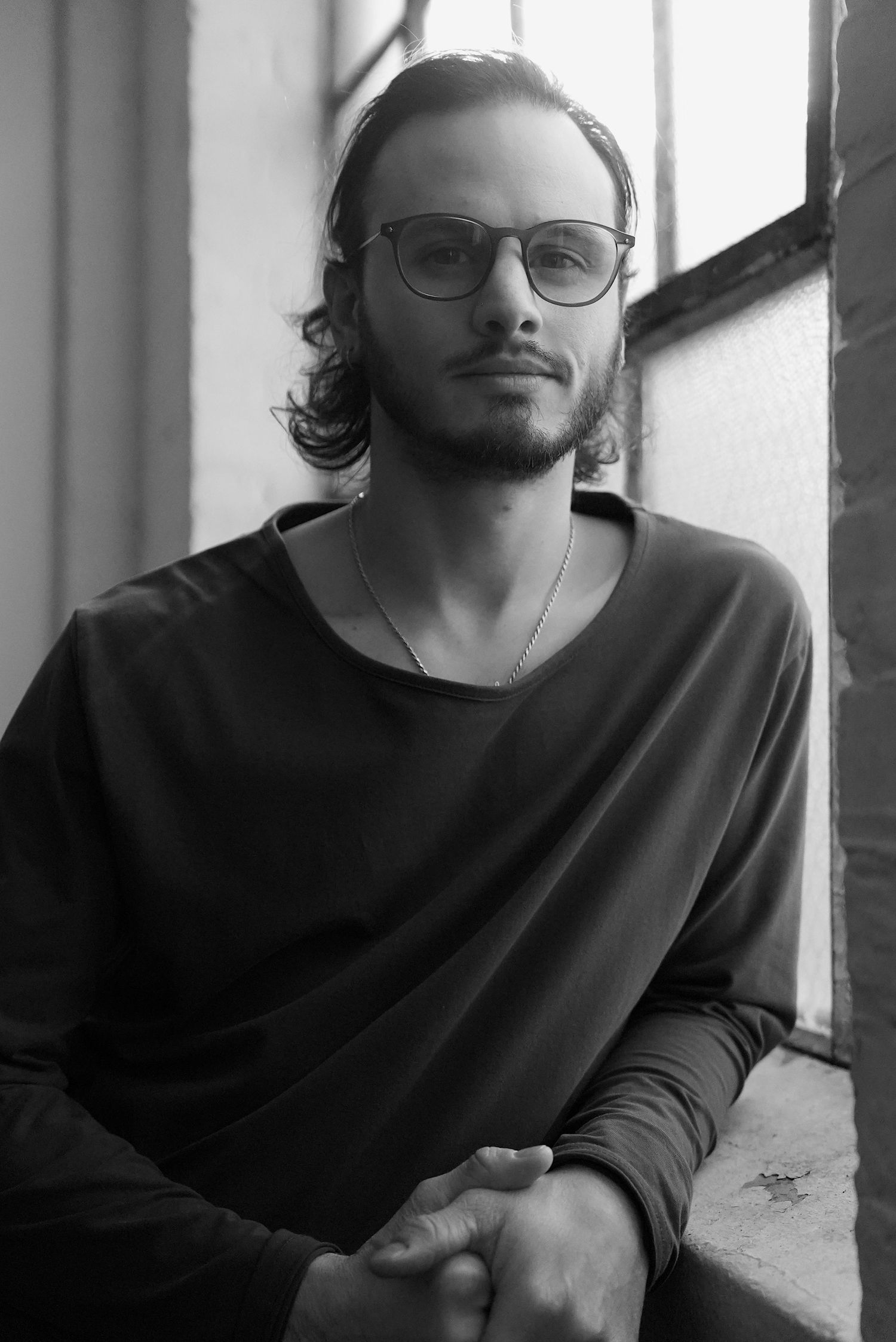 Black and white portrait of young man with glasses and wavy hair sitting by window.