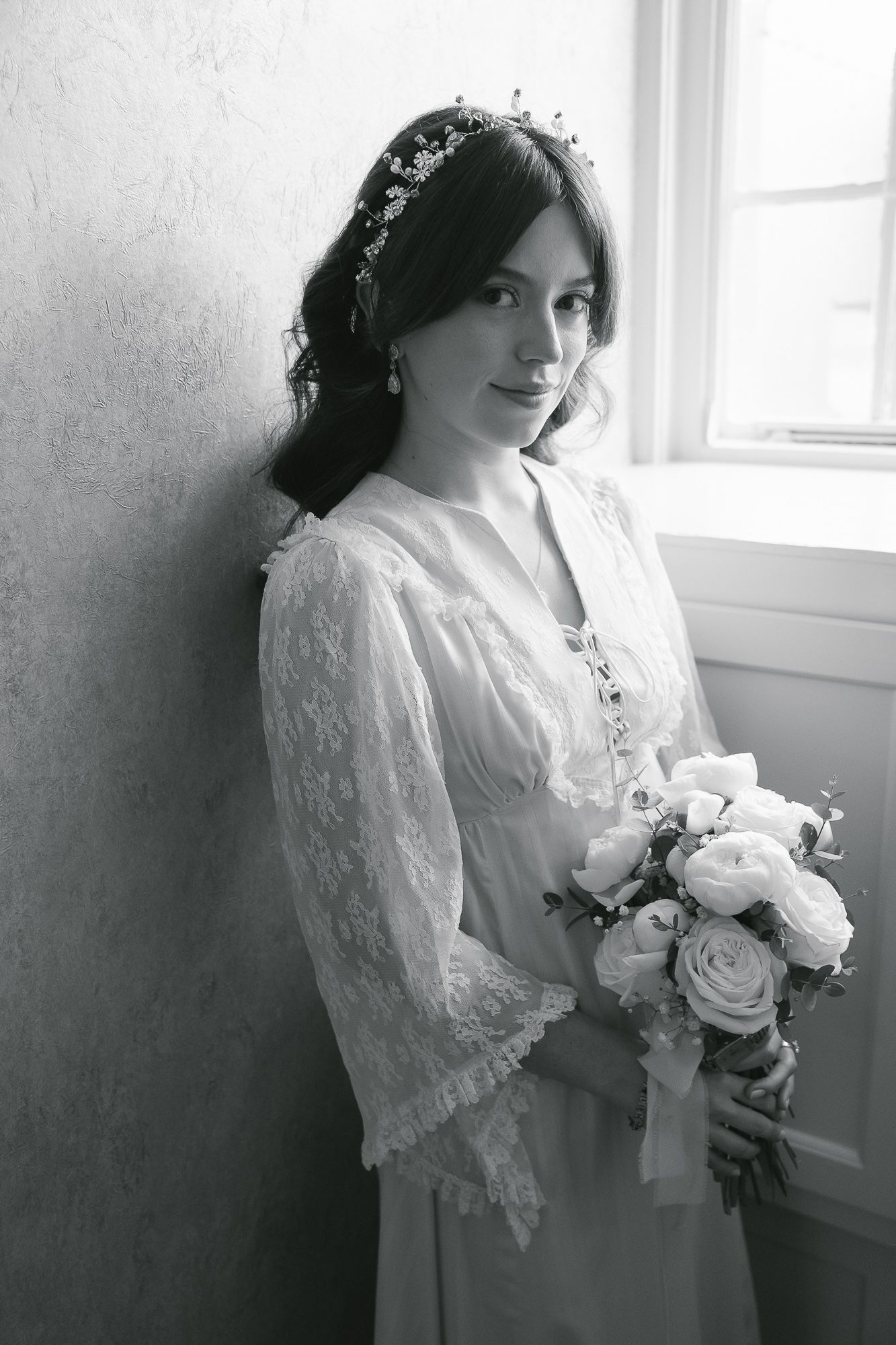 Elegant bride in vintage lace gown holds white bouquet while leaning against wall in B&W photo.