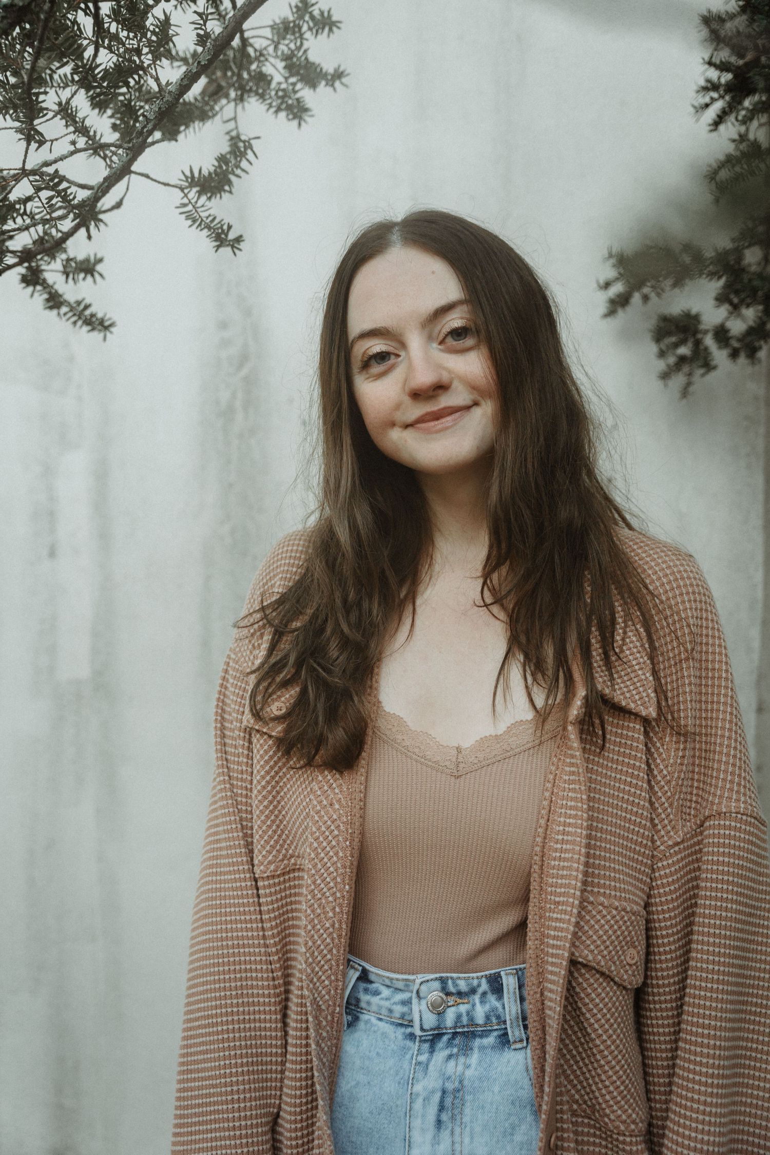 A young brunette woman in a tan cardigan and jeans smiles softly against a light grey wall.
