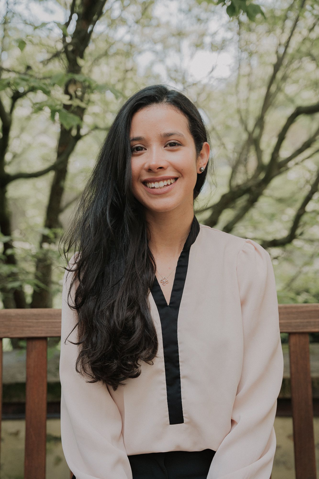 Smiling young woman with long dark hair in cream blouse with black trim posing outdoors near wooden fence.