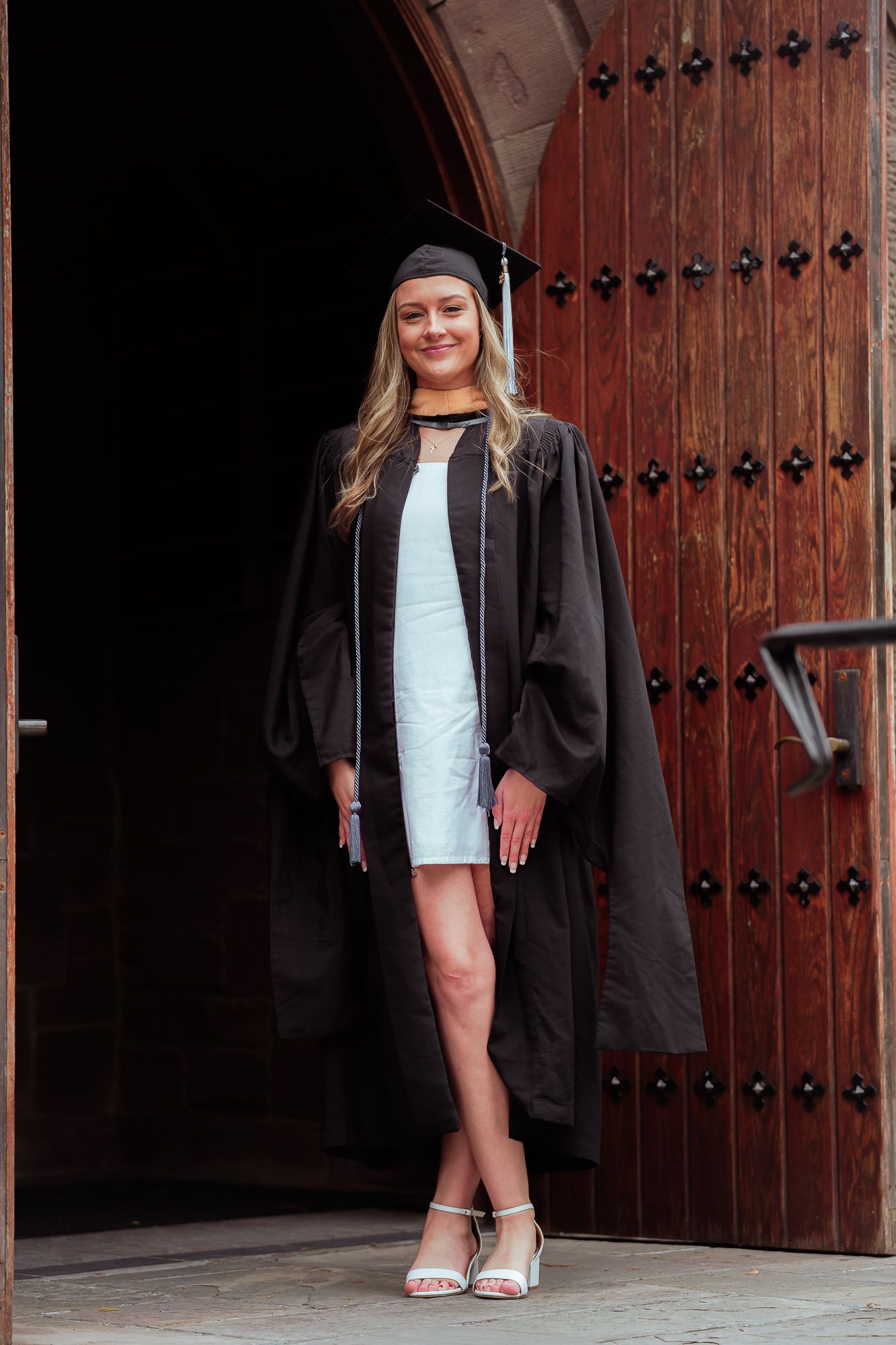 Smiling female graduate in black cap and gown posing in front of large wooden arched doors.
