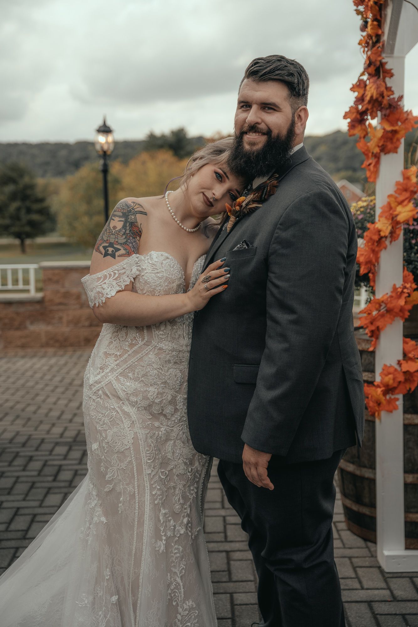 Bride in lace off-shoulder gown leans on bearded groom in dark suit at fall outdoor wedding ceremony.