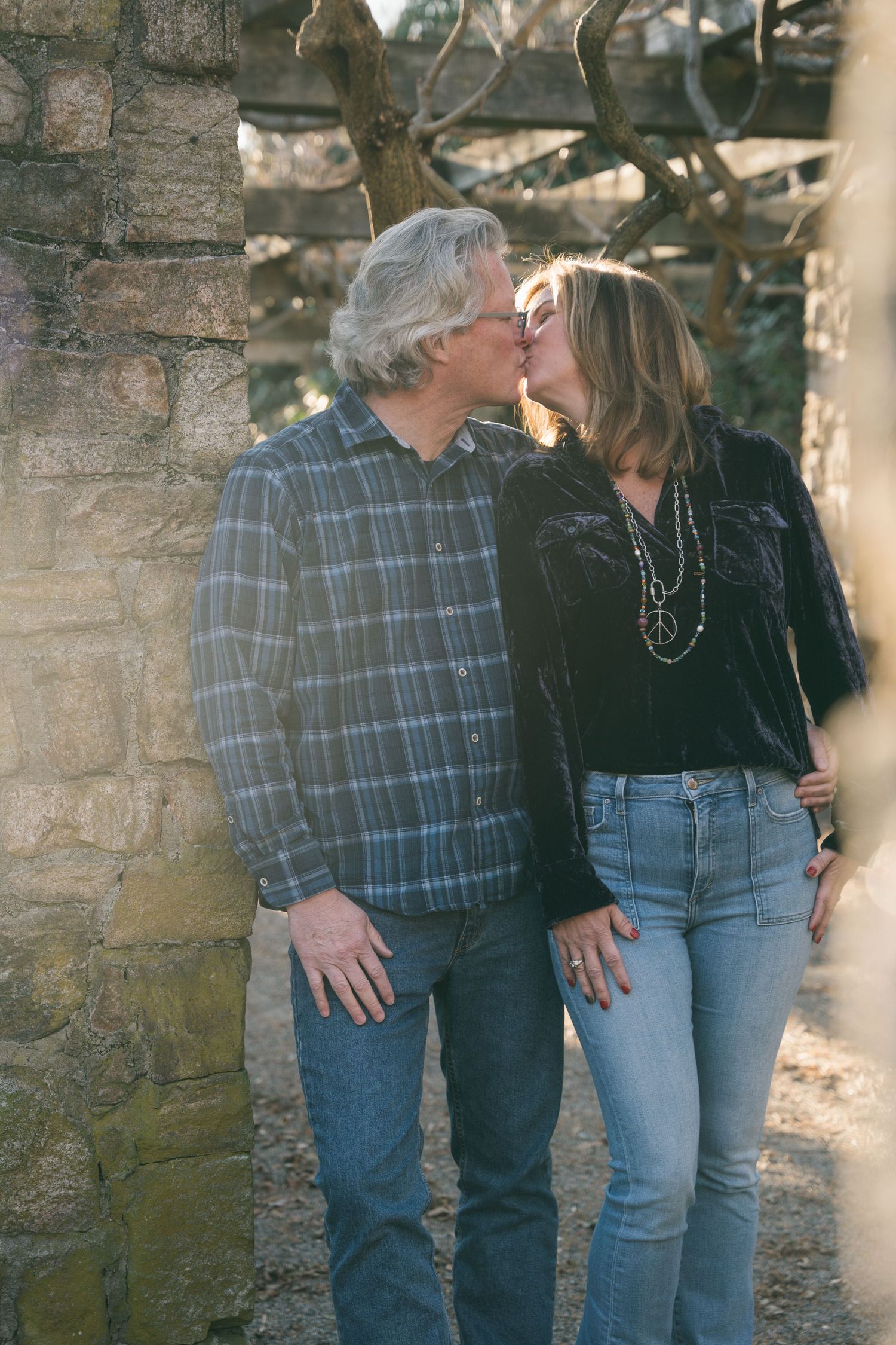 Mature couple sharing a kiss outdoors near a stone wall in casual jeans and plaid shirt.