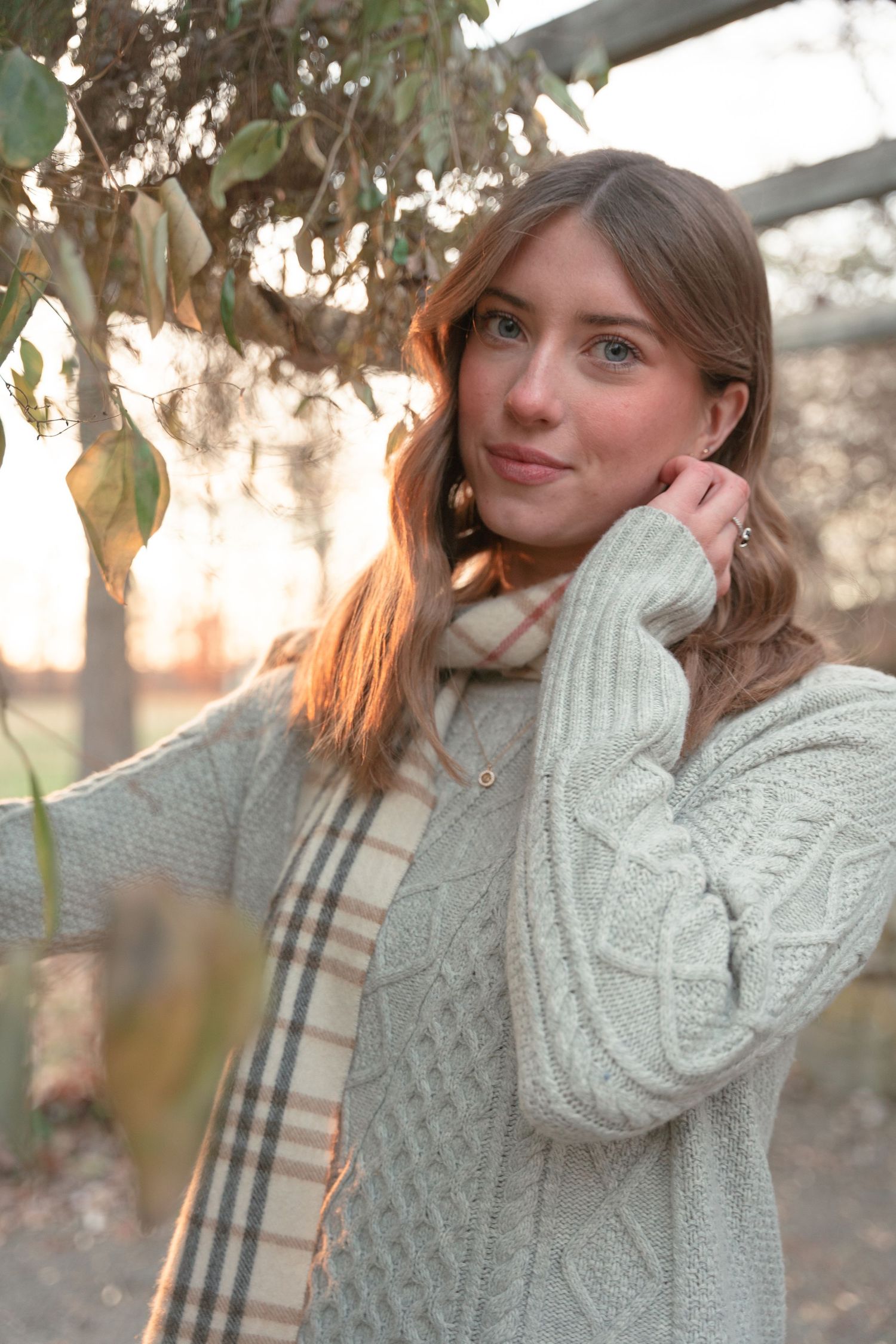 Young woman in cream cable knit sweater and plaid scarf posing outdoors in golden sunset light.