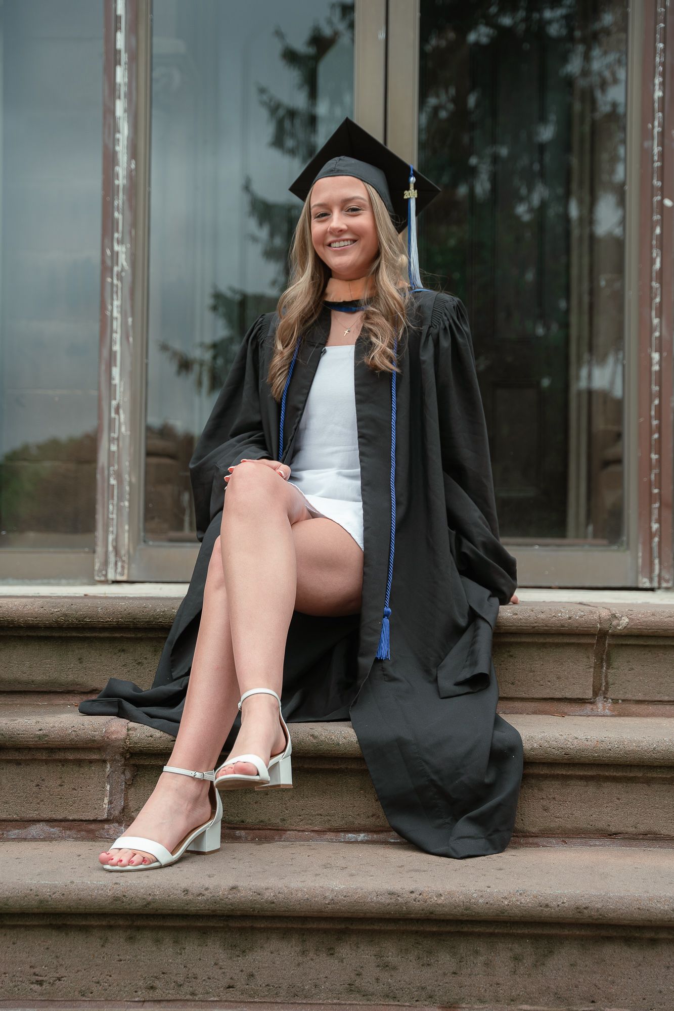 Happy blonde graduate in cap and gown sits on stone steps showing off white heels and dress.