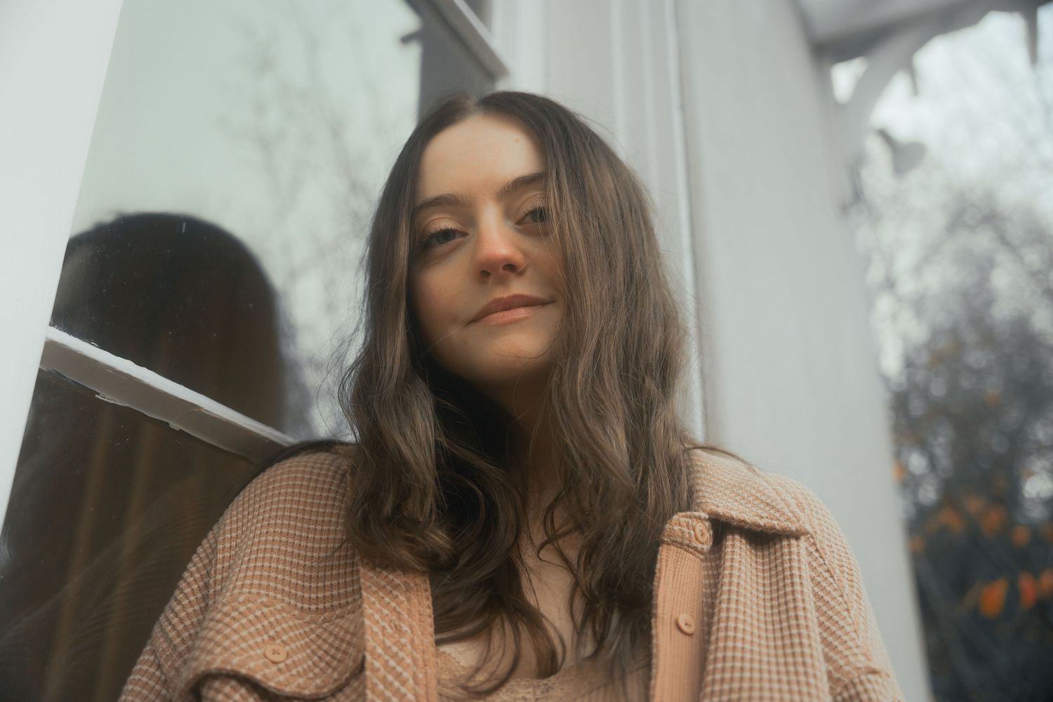 Young woman with long brown hair wearing a beige coat poses by a window in soft natural light.