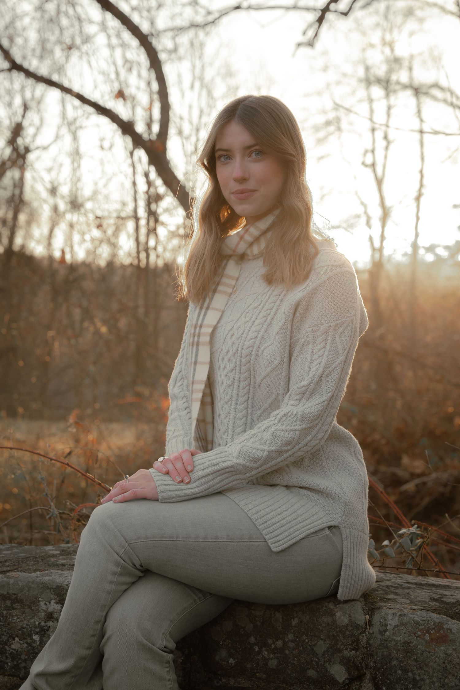 Young woman in cream knit sweater sitting on a rock in a sunlit autumn outdoor setting.