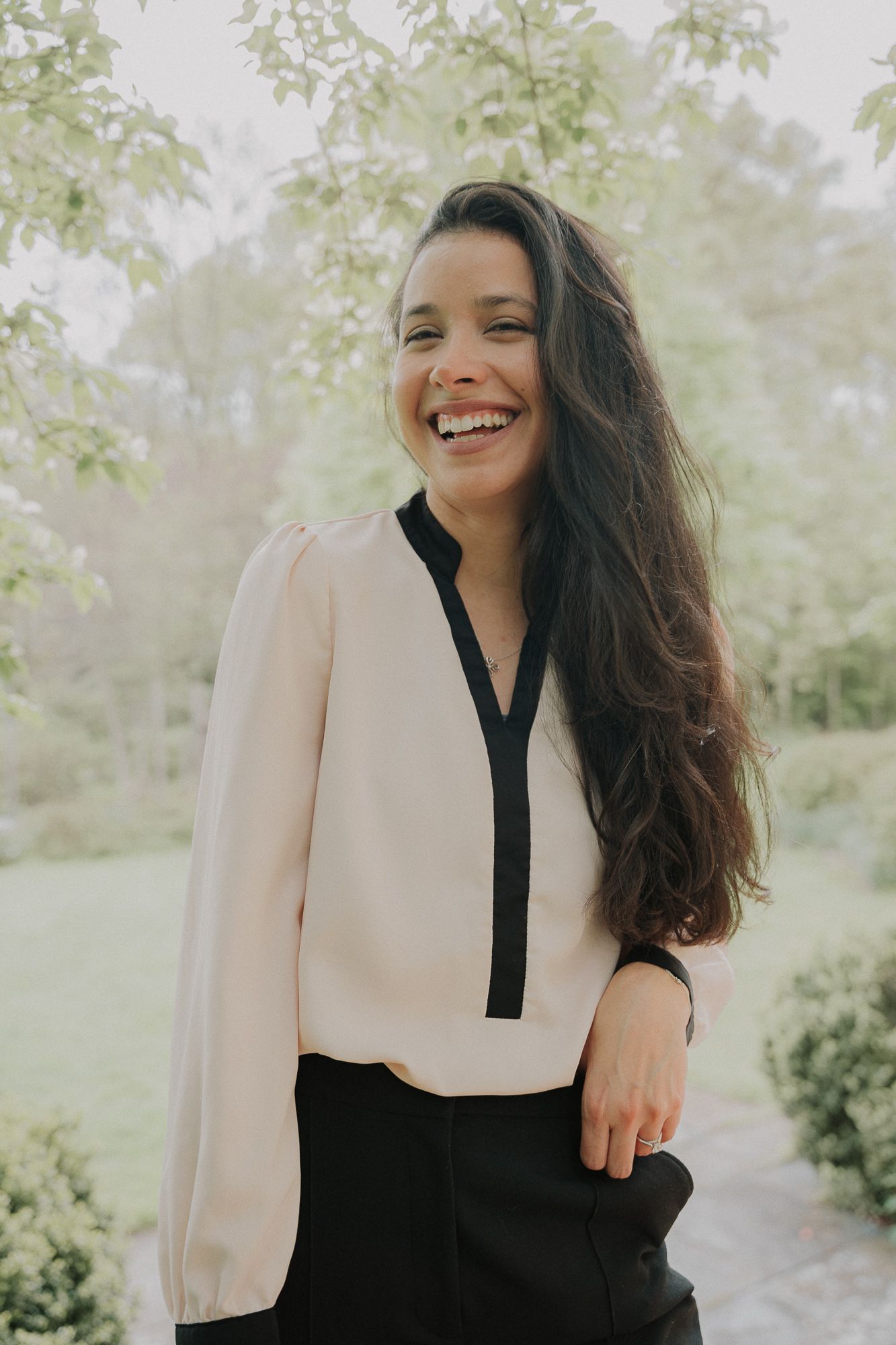 Laughing young woman with long dark hair wearing cream and black blouse outdoors in garden setting.
