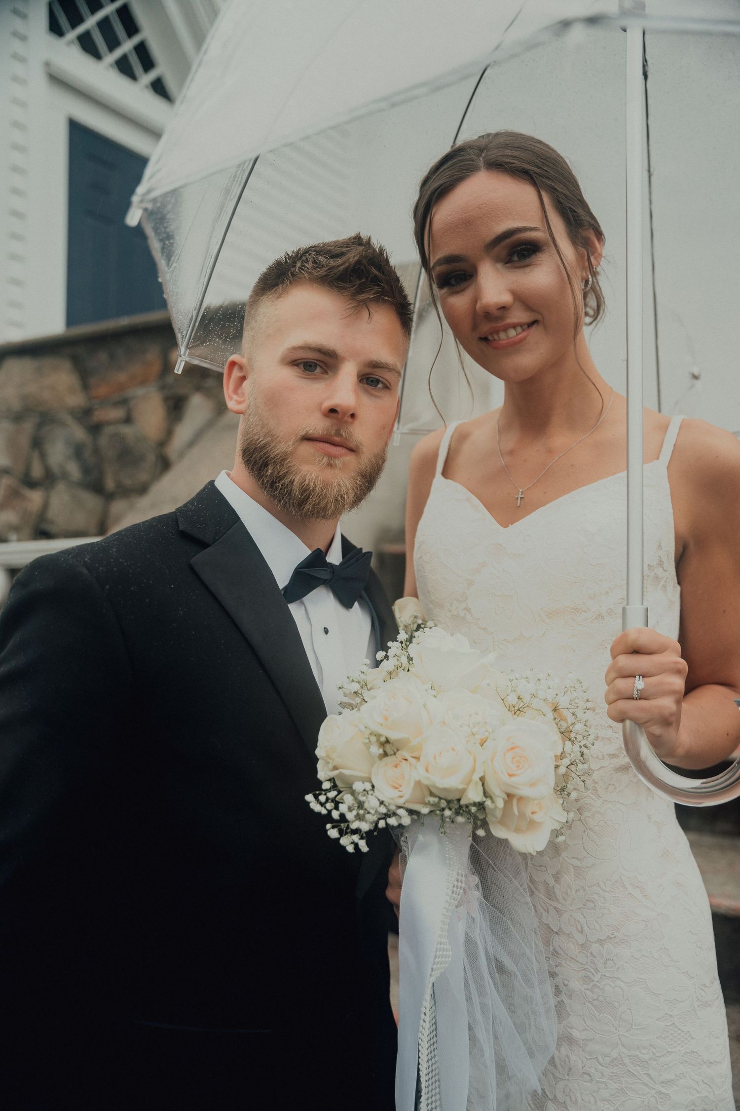 Bride and groom pose under a clear umbrella on their rainy wedding day holding white bouquet.