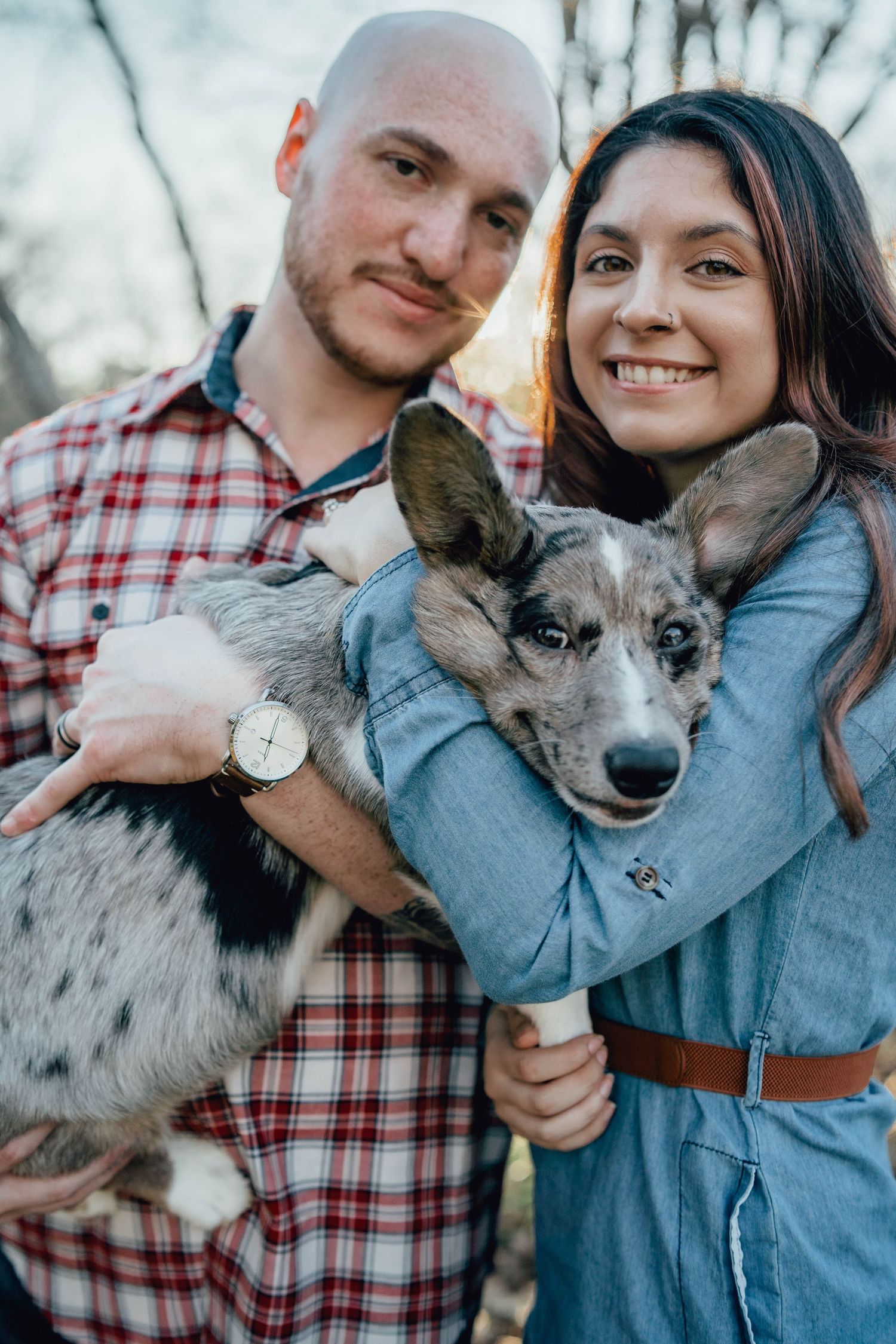 A smiling couple holds an adorable merle puppy outdoors in warm golden hour light.