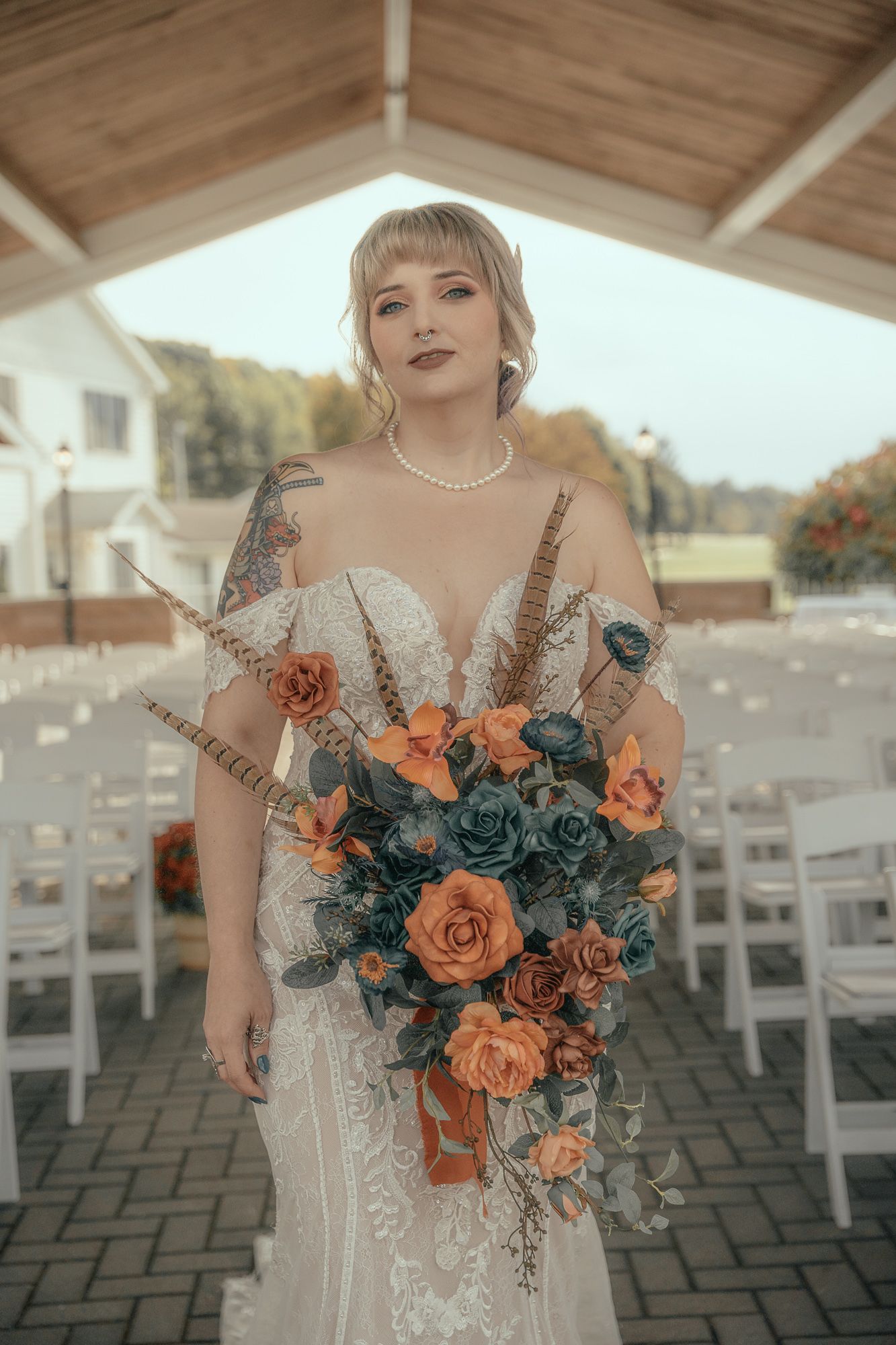 Bold bride with tattoos and blonde bangs holds large orange and teal bouquet at outdoor ceremony.