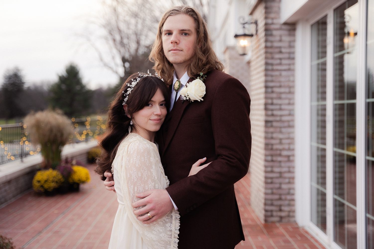 Bride in lace gown leans against groom in burgundy suit on outdoor patio during wedding.