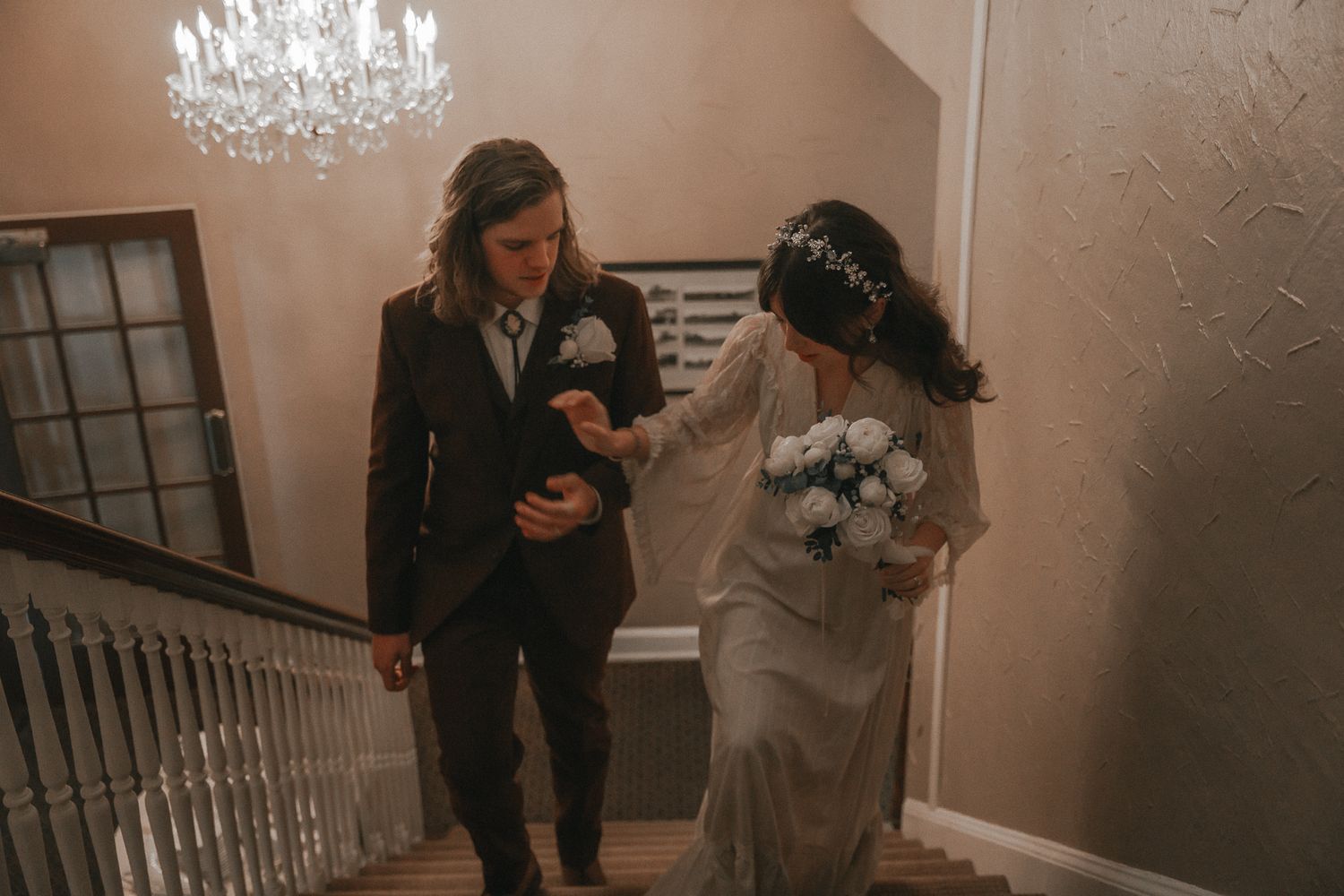 Bride and groom descend staircase together under chandelier as bride carries blue floral bouquet.
