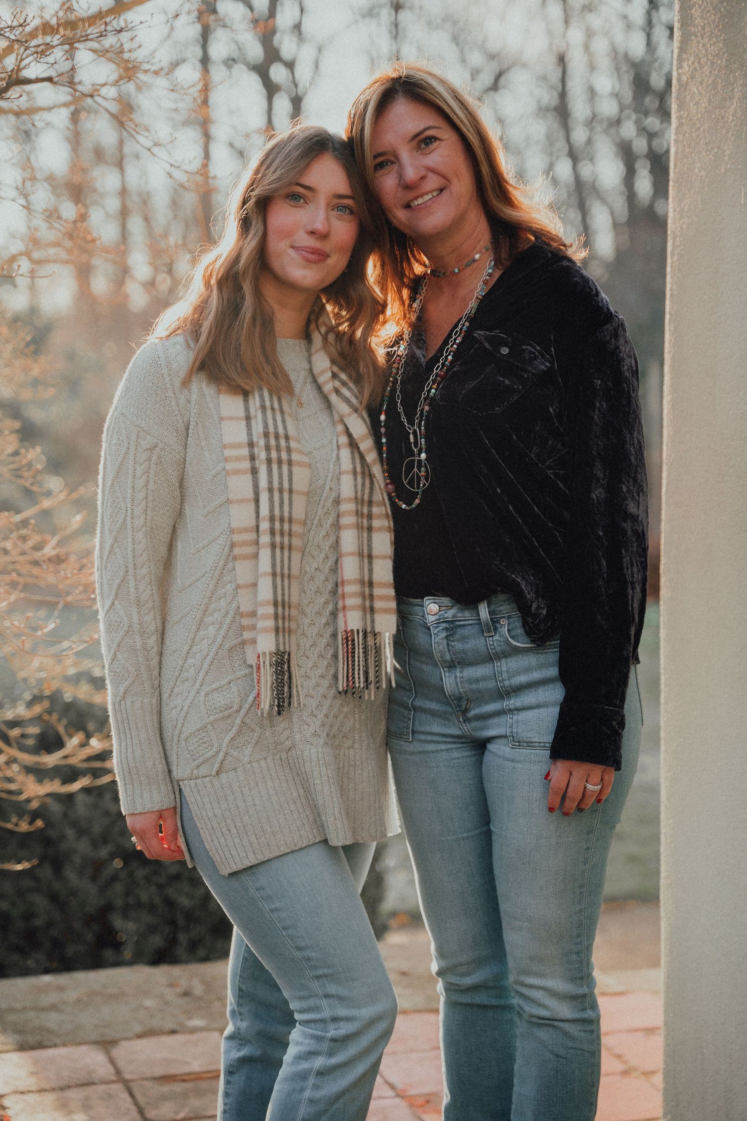 Mother and daughter smiling together outdoors at sunset, wearing jeans and cozy sweaters.