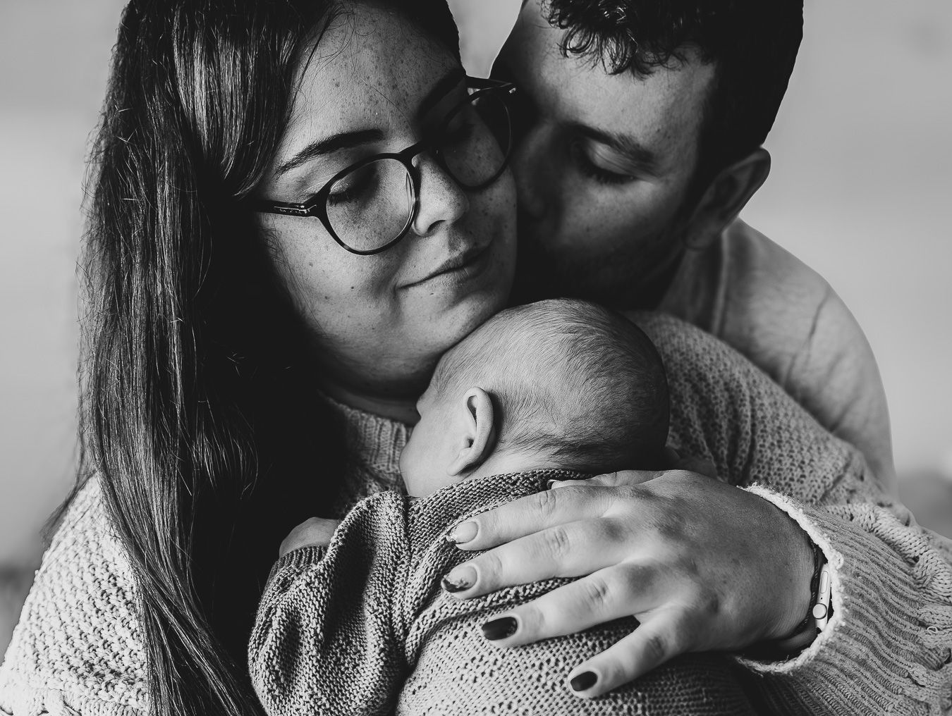 Parents tenderly embracing their newborn baby in a black and white family portrait.