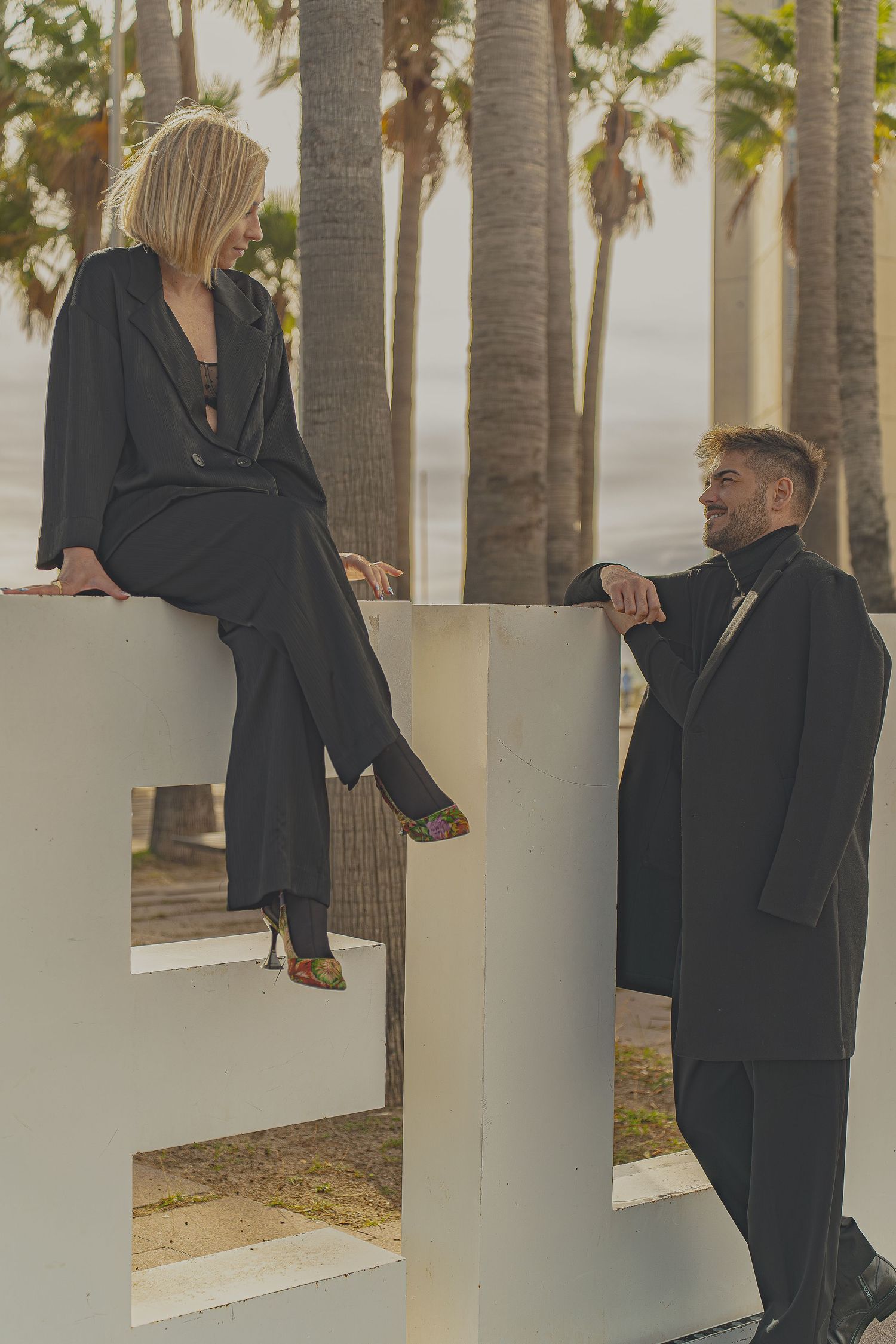 A blonde woman and a man in black suits pose near large white letters with palm trees and ocean in the background.