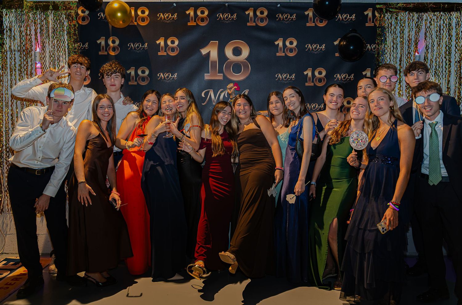 Group of teens in formal attire posing in front of an 18th birthday party backdrop with gold decorations.