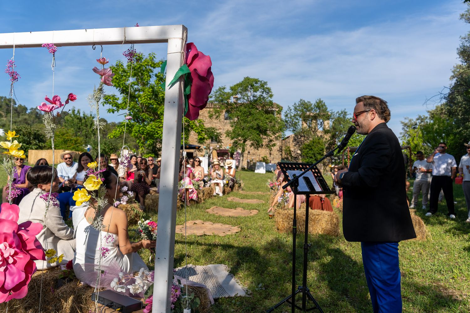 Outdoor wedding ceremony with guests seated on grass, officiant at podium, colorful floral arch decoration.