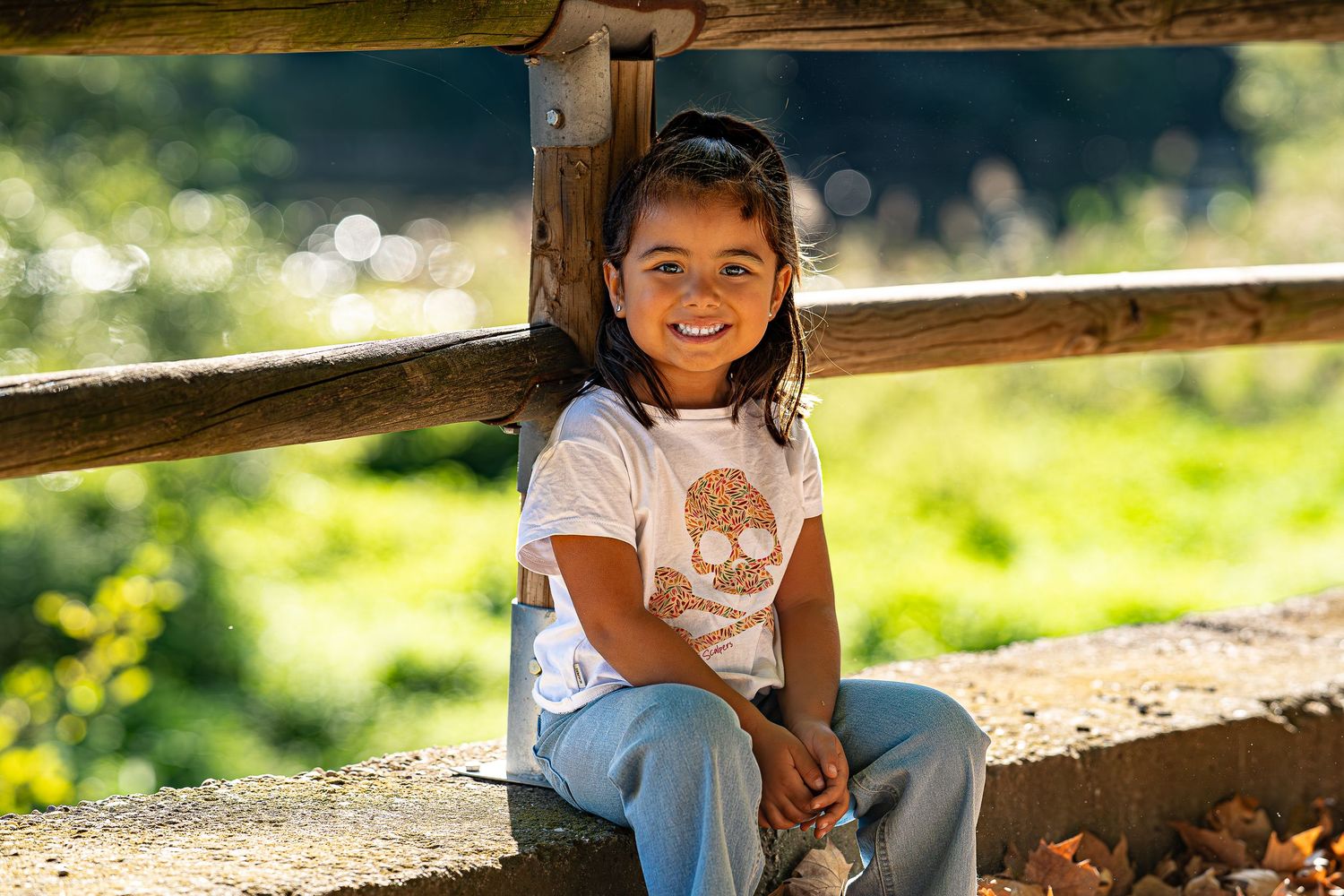 Smiling young girl in white t-shirt and blue pants sitting on a wooden fence in a sunny green outdoor setting.