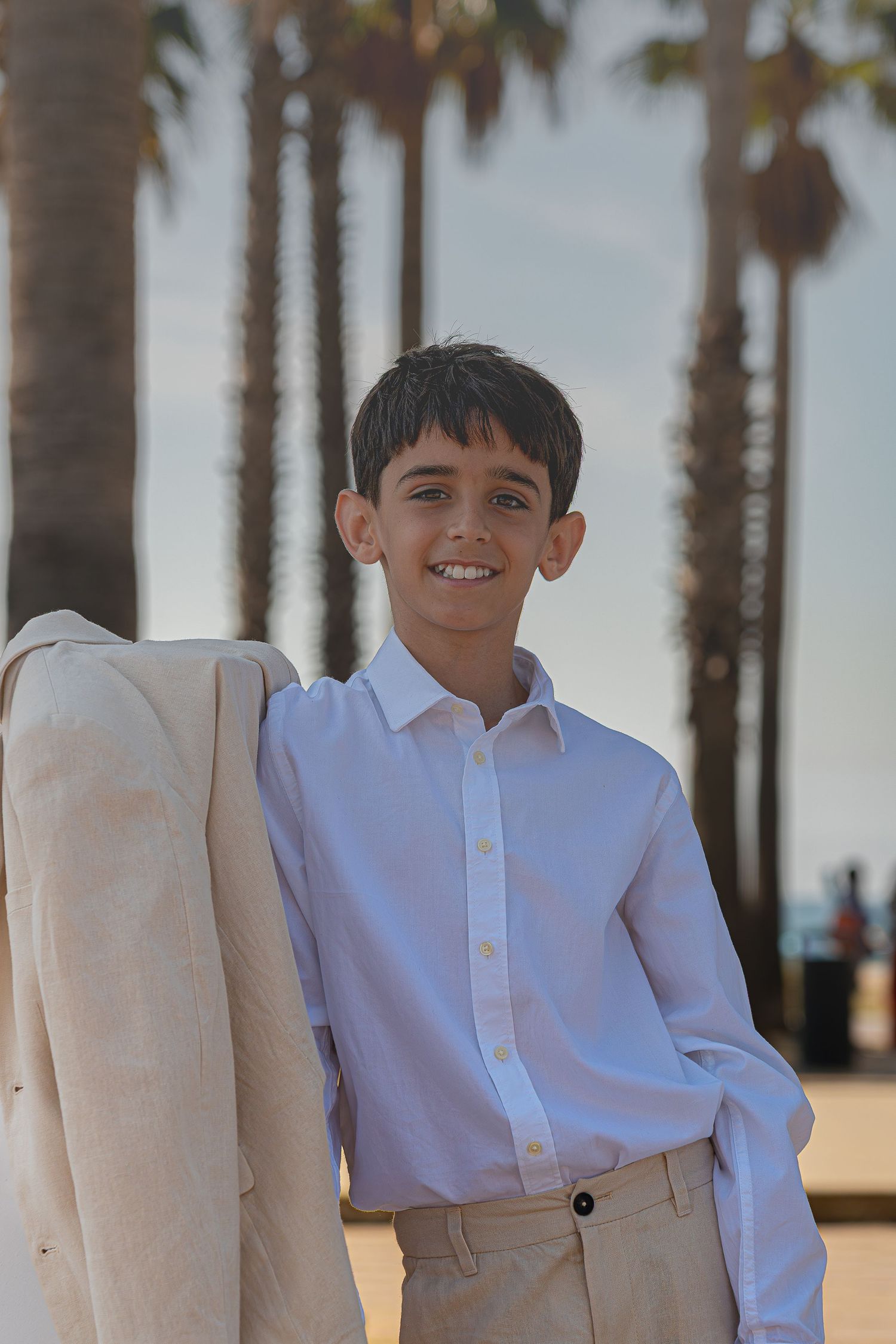 Smiling boy in white shirt holding beige jacket with palm trees in background at sunset.