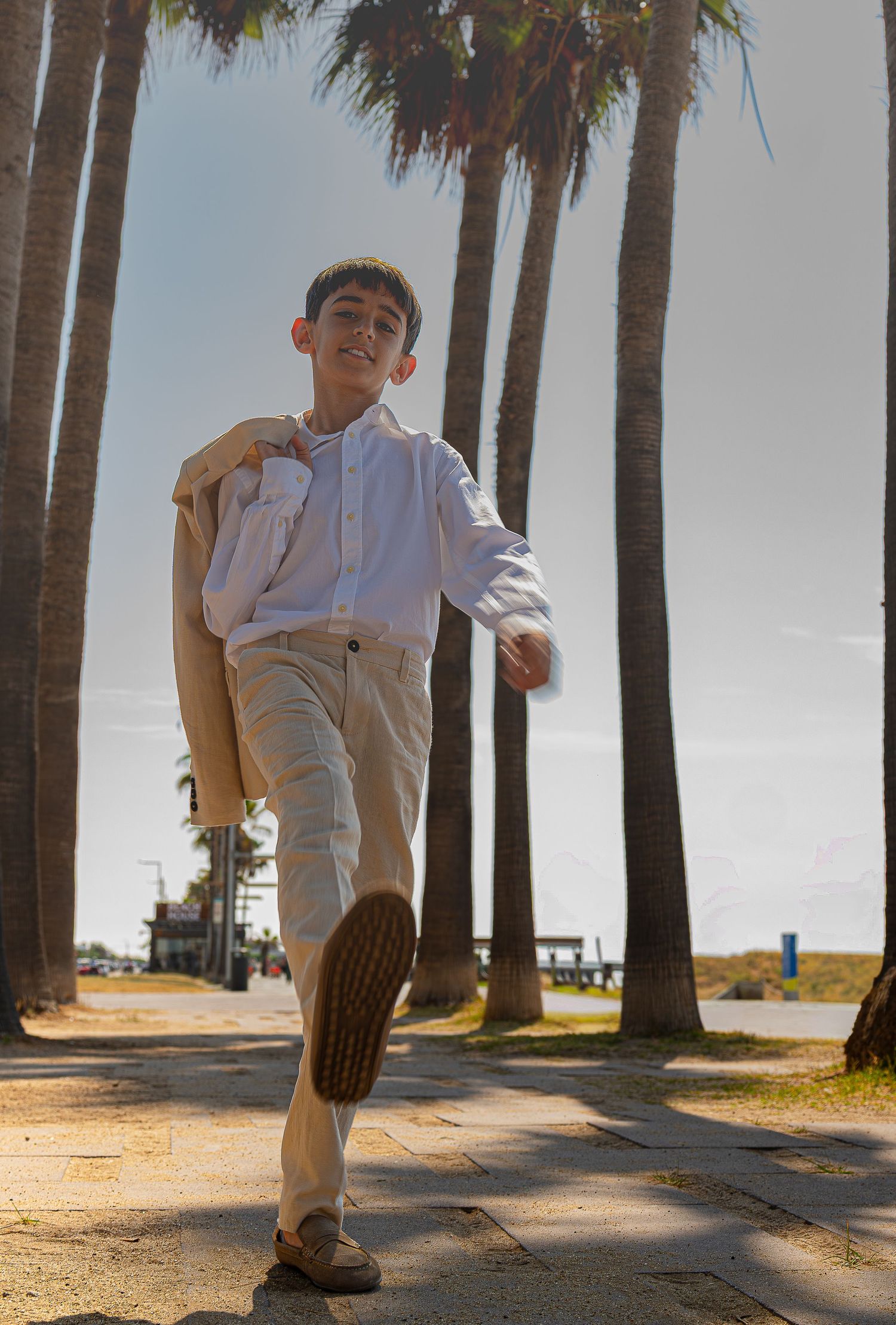 A smiling boy walks confidently along a palm-lined path, wearing a white shirt and beige pants on a sunny day.