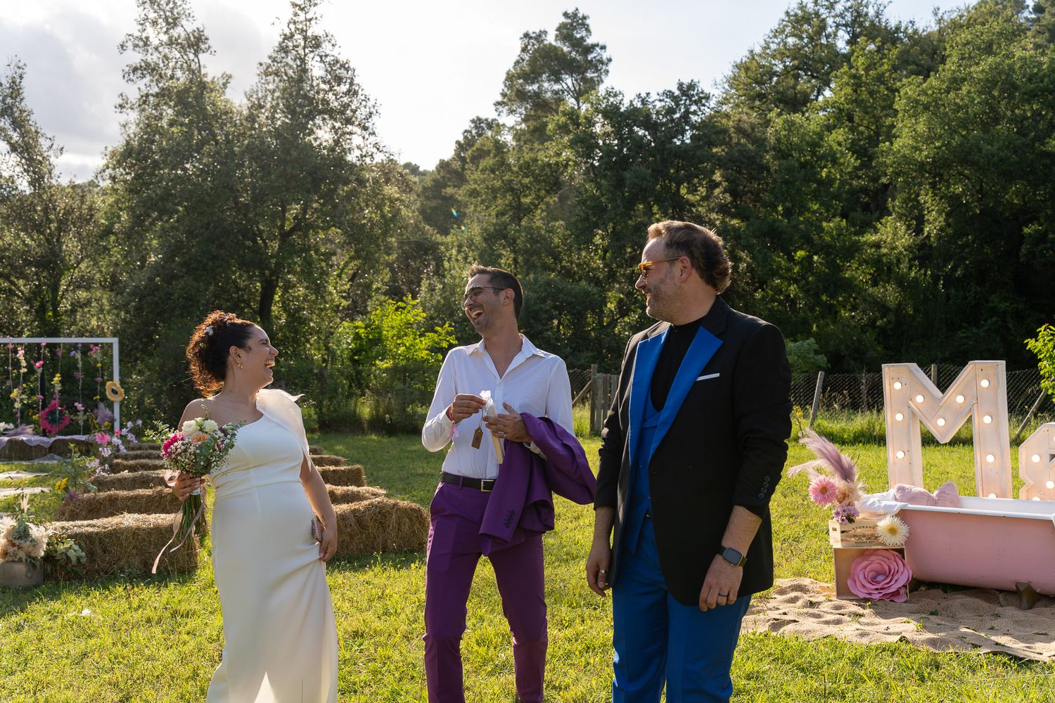 Three people in colorful formal attire chat outdoors at a garden wedding ceremony with hay bales and pink decorations.