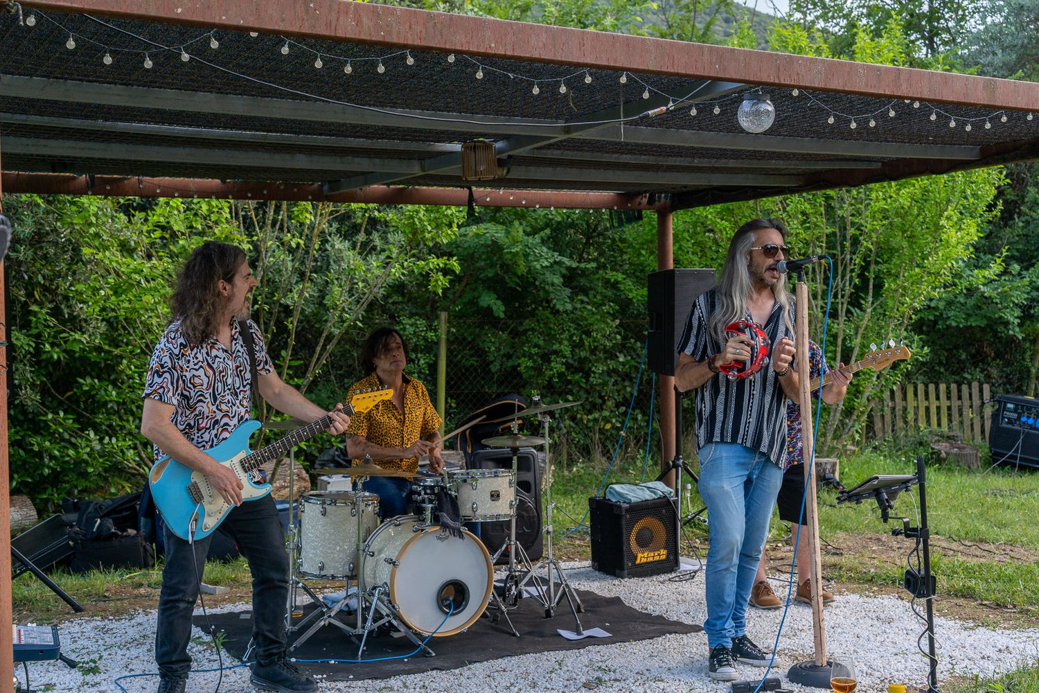 Outdoor band performing live music under a covered stage with guitarist, drummer, and vocalist in a lush green backyard setting.