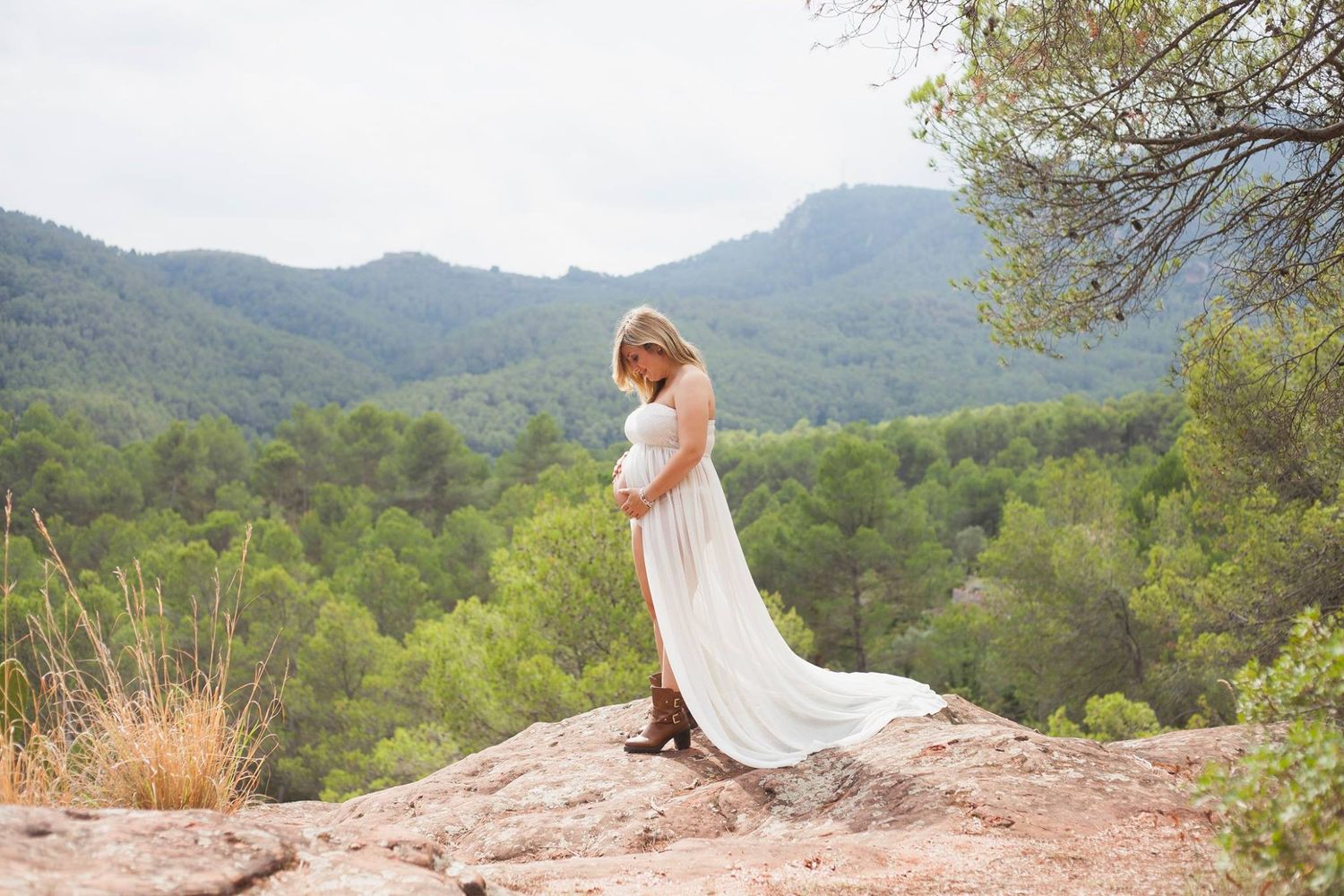 Mujer con un vestido blanco vaporoso posa con elegancia en un acantilado rocoso con vistas a un pintoresco paisaje montañoso.