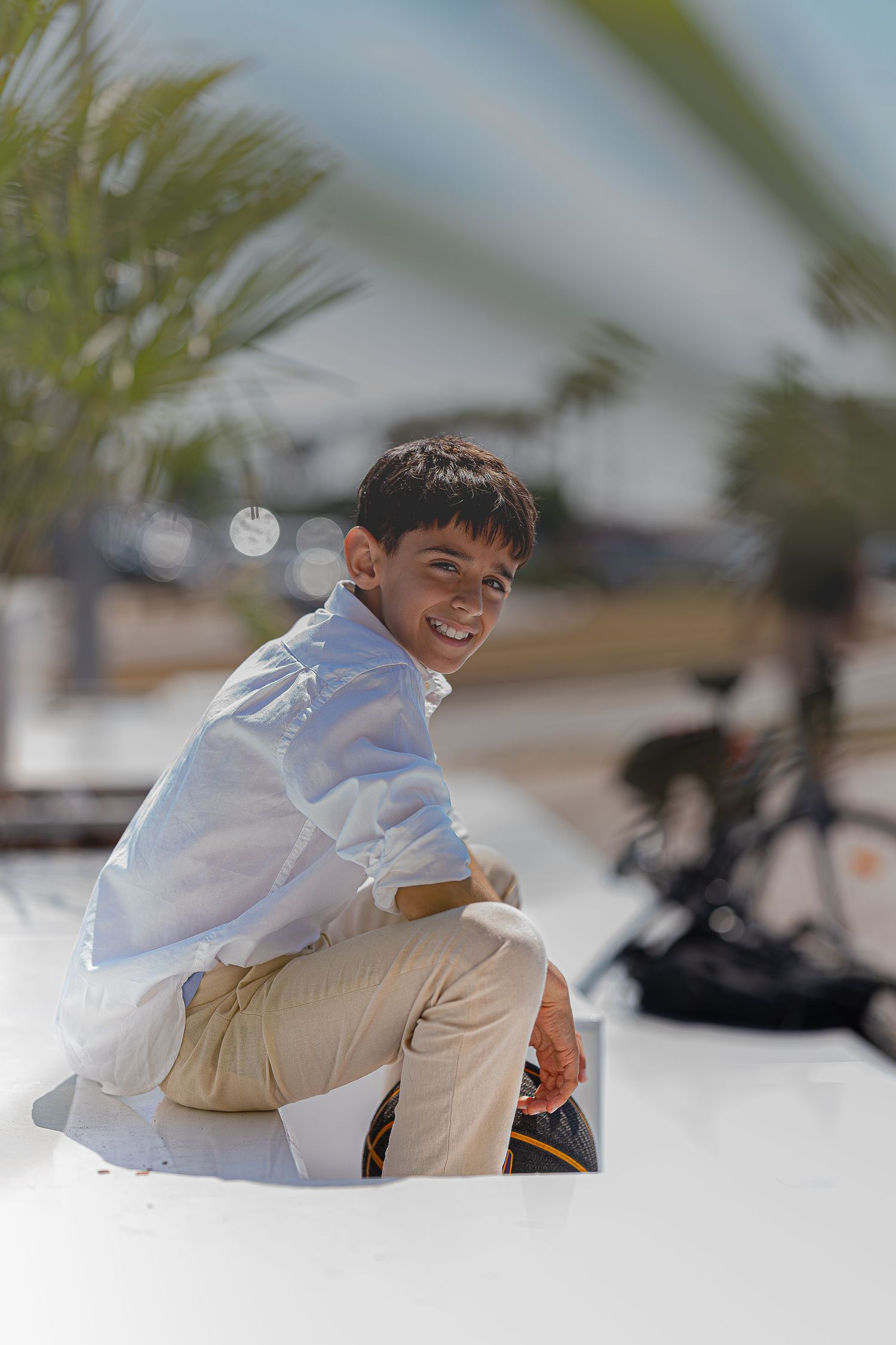 Happy boy in white shirt sitting outdoors near palm trees and a bicycle on a sunny day.
