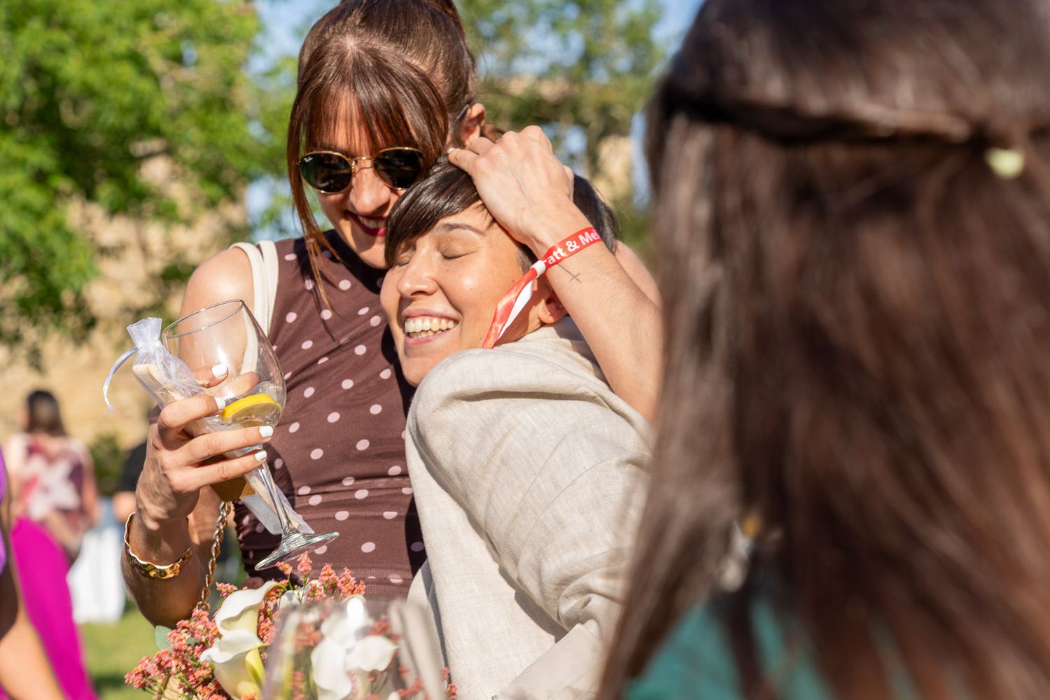 Two women laughing and embracing outdoors at a sunny social gathering, one holding a drink.