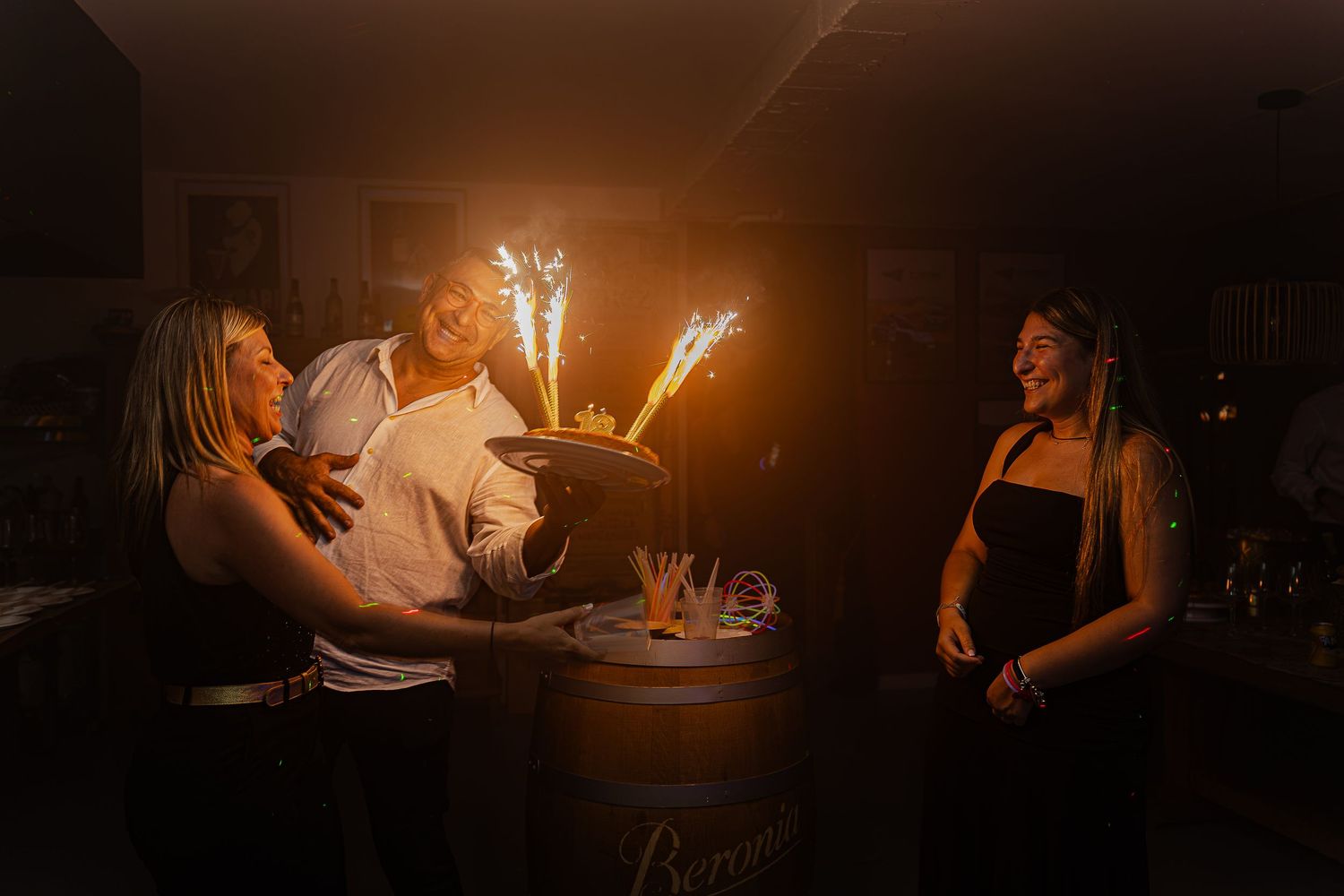 People celebrating with sparkler-topped dessert on a wine barrel in a dimly lit rustic setting.