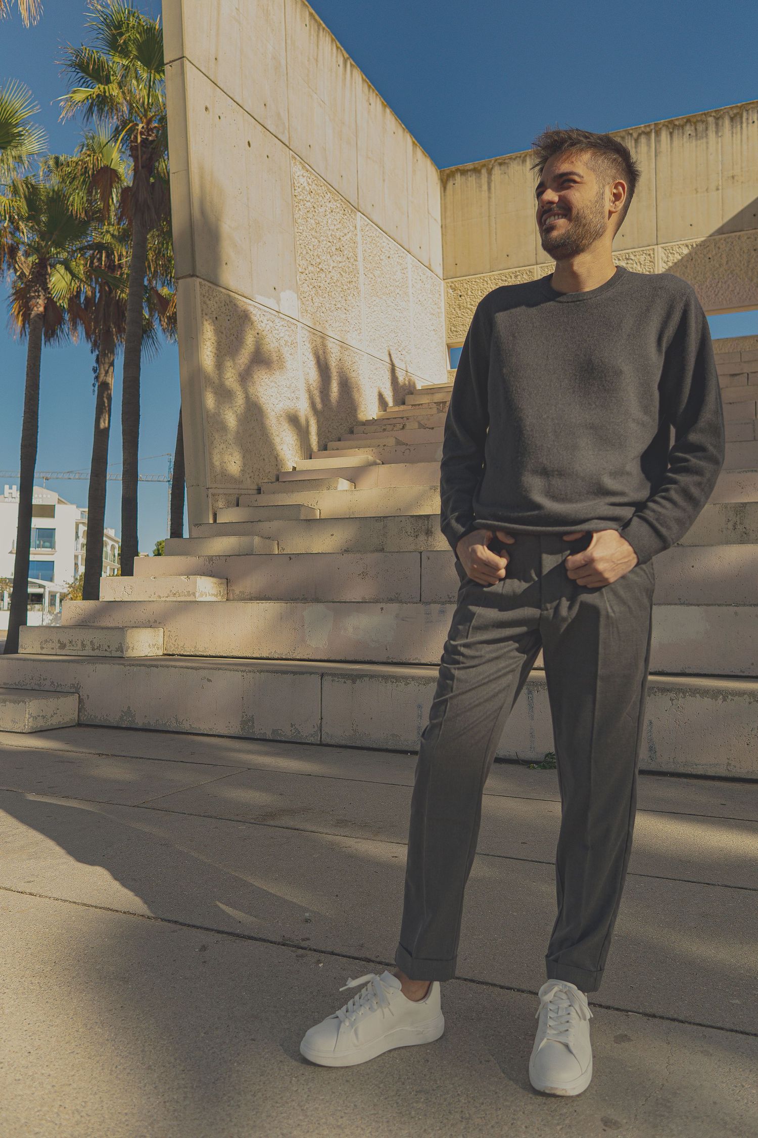 Man in dark sweatshirt and gray pants poses by concrete steps with palm trees under blue sky.