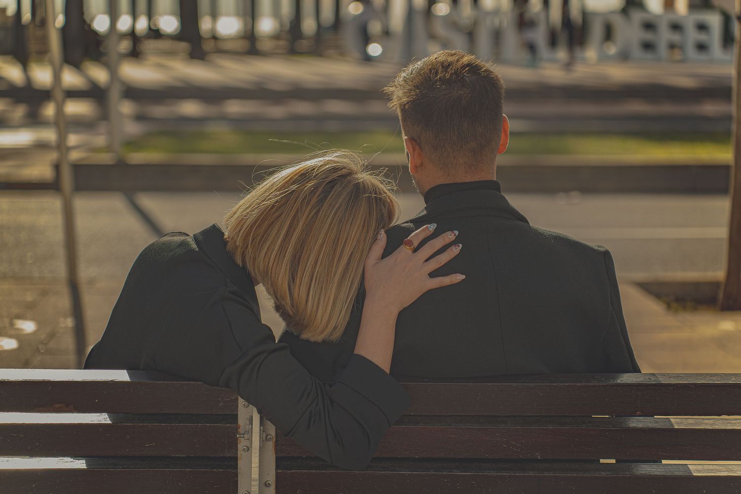 Couple sitting on a bench from behind, woman resting head on man's shoulder in a romantic outdoor setting.