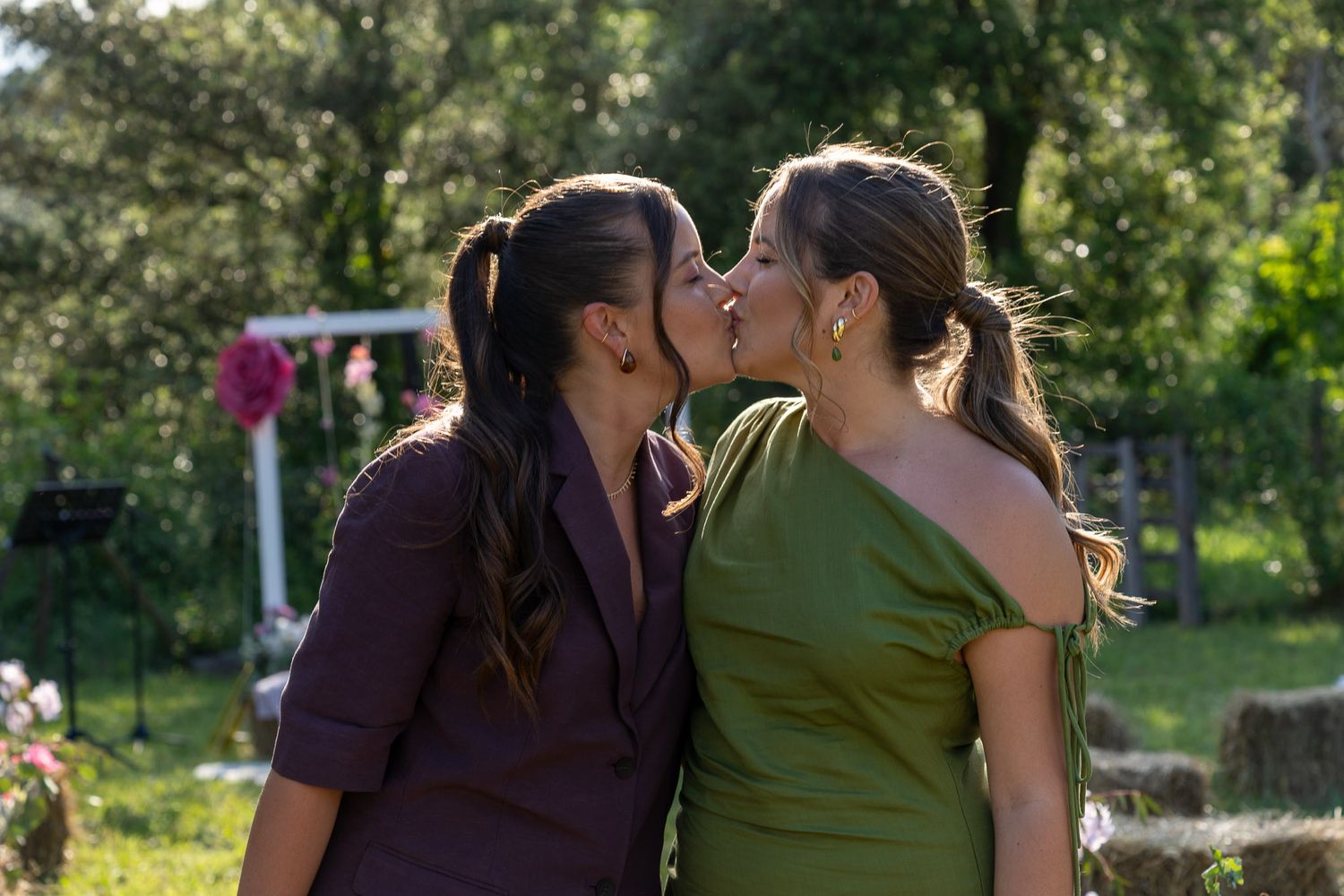 Two women in elegant dresses sharing a kiss outdoors at a garden celebration.