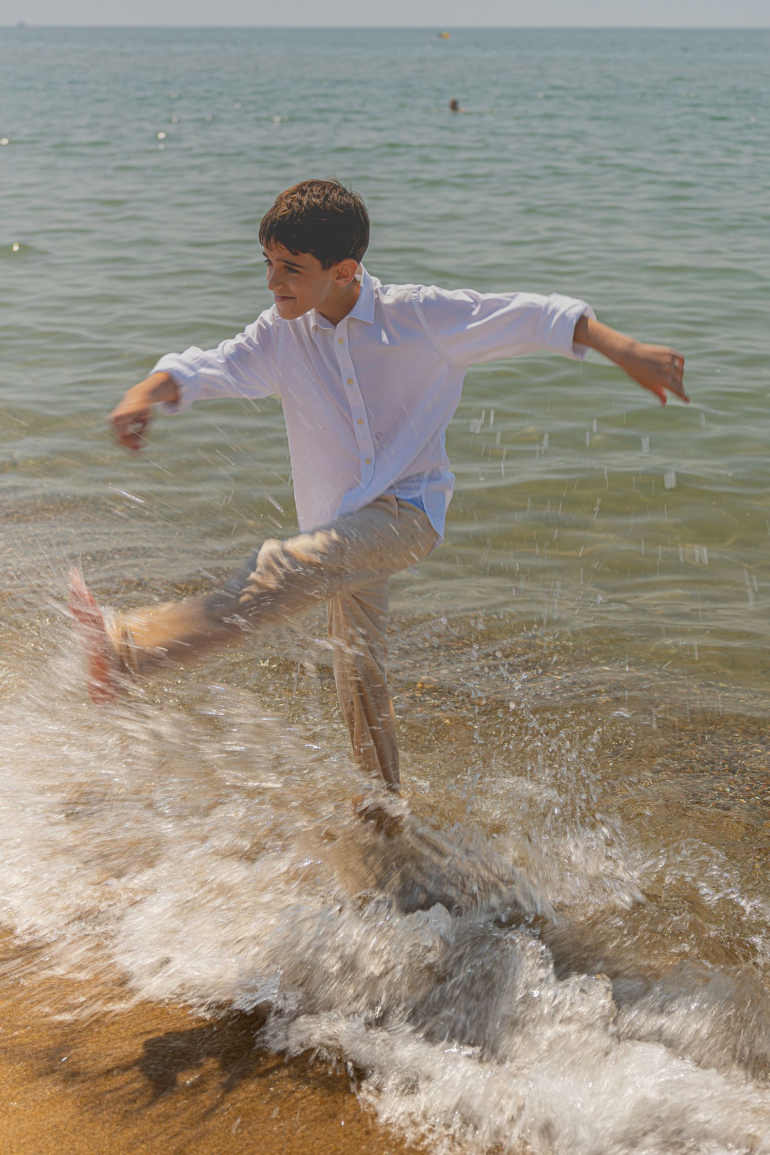 A young boy in a white shirt splashing through ocean waves on a sandy beach on a sunny day.