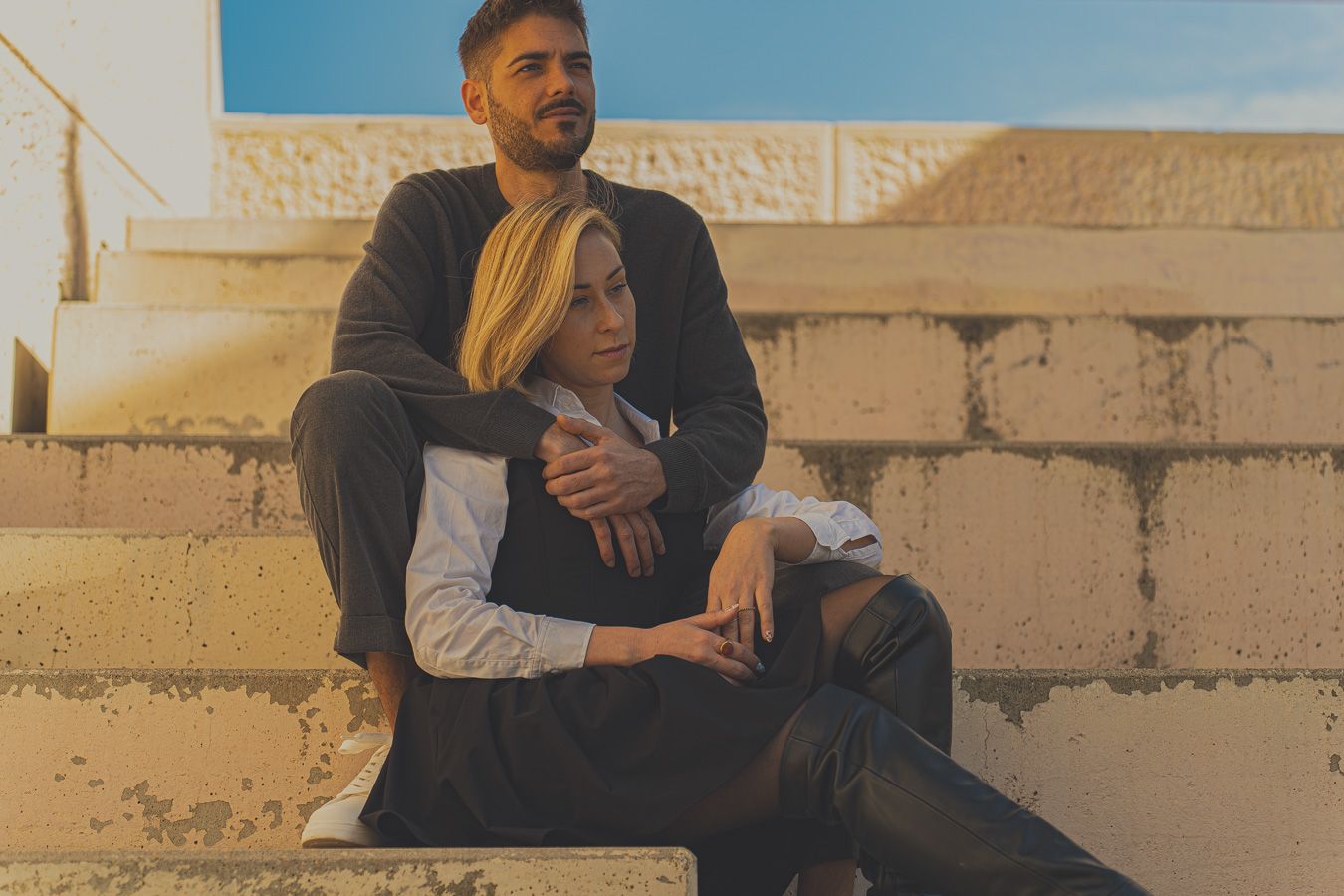 Pareja sentada y abrazada en unos escalones de piedra con una pared iluminada por el sol al fondo, vestida con ropa informal.