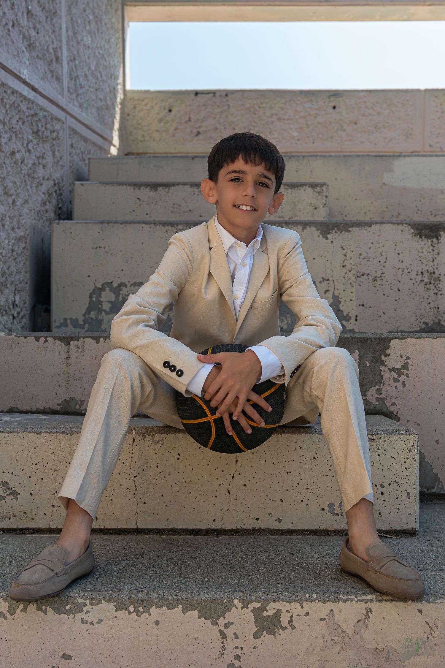 Young boy in beige suit sitting on concrete steps holding a basketball, smiling at camera.