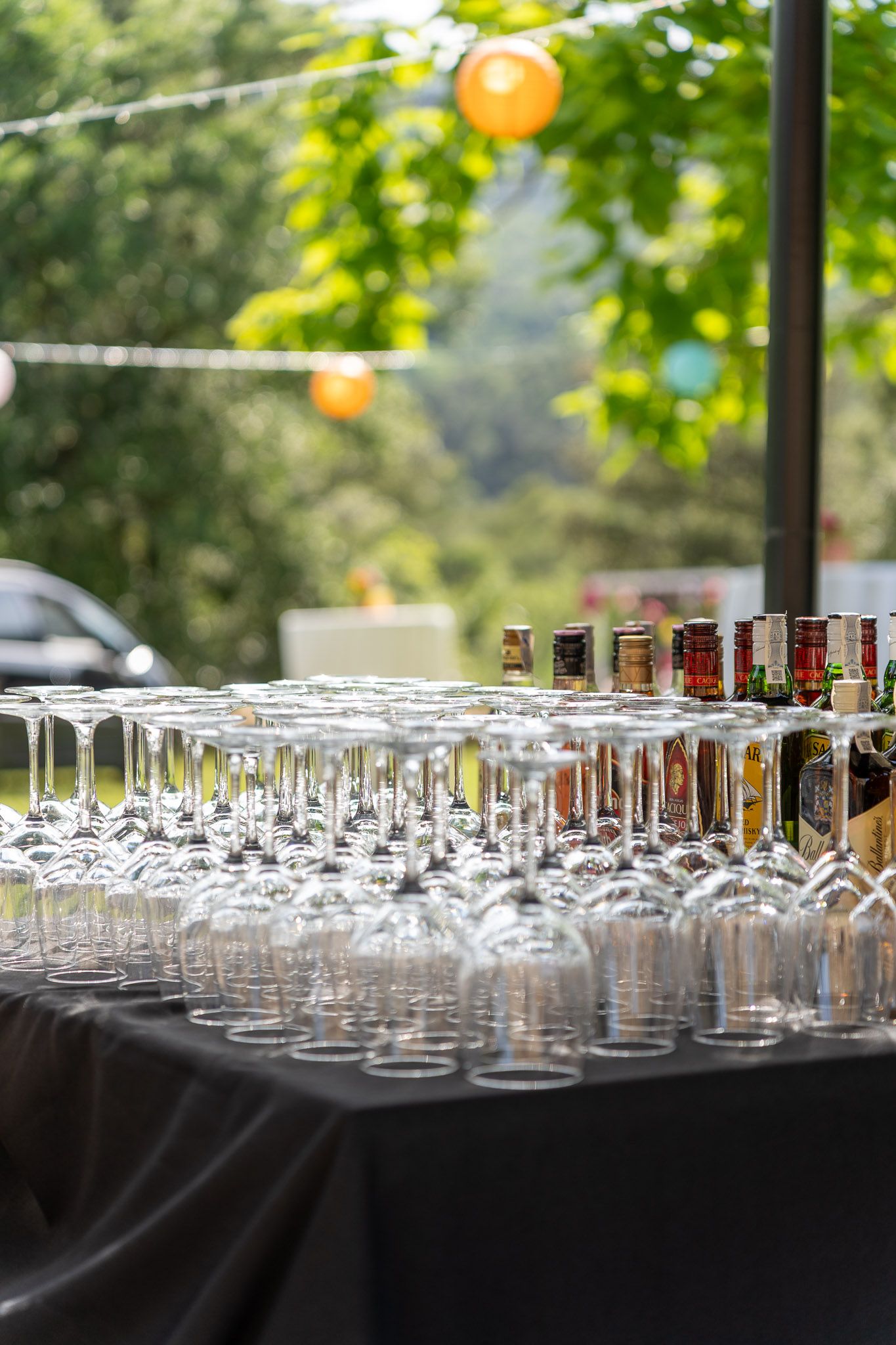 Rows of empty wine glasses and liquor bottles arranged on a black draped table at an outdoor party event.