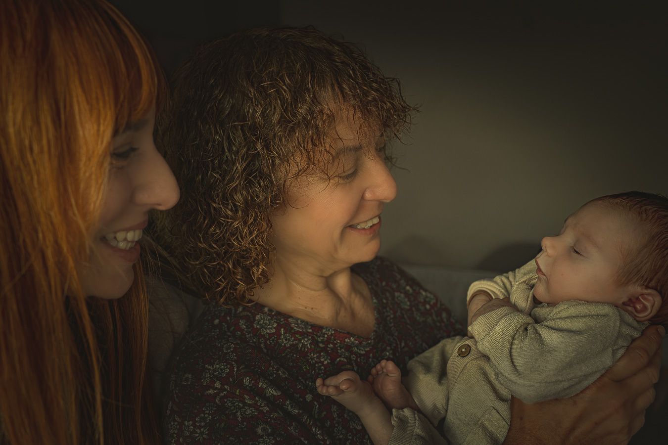 Two women smiling at a newborn baby being cradled, one with red hair and one with curly hair, in a warm indoor setting.
