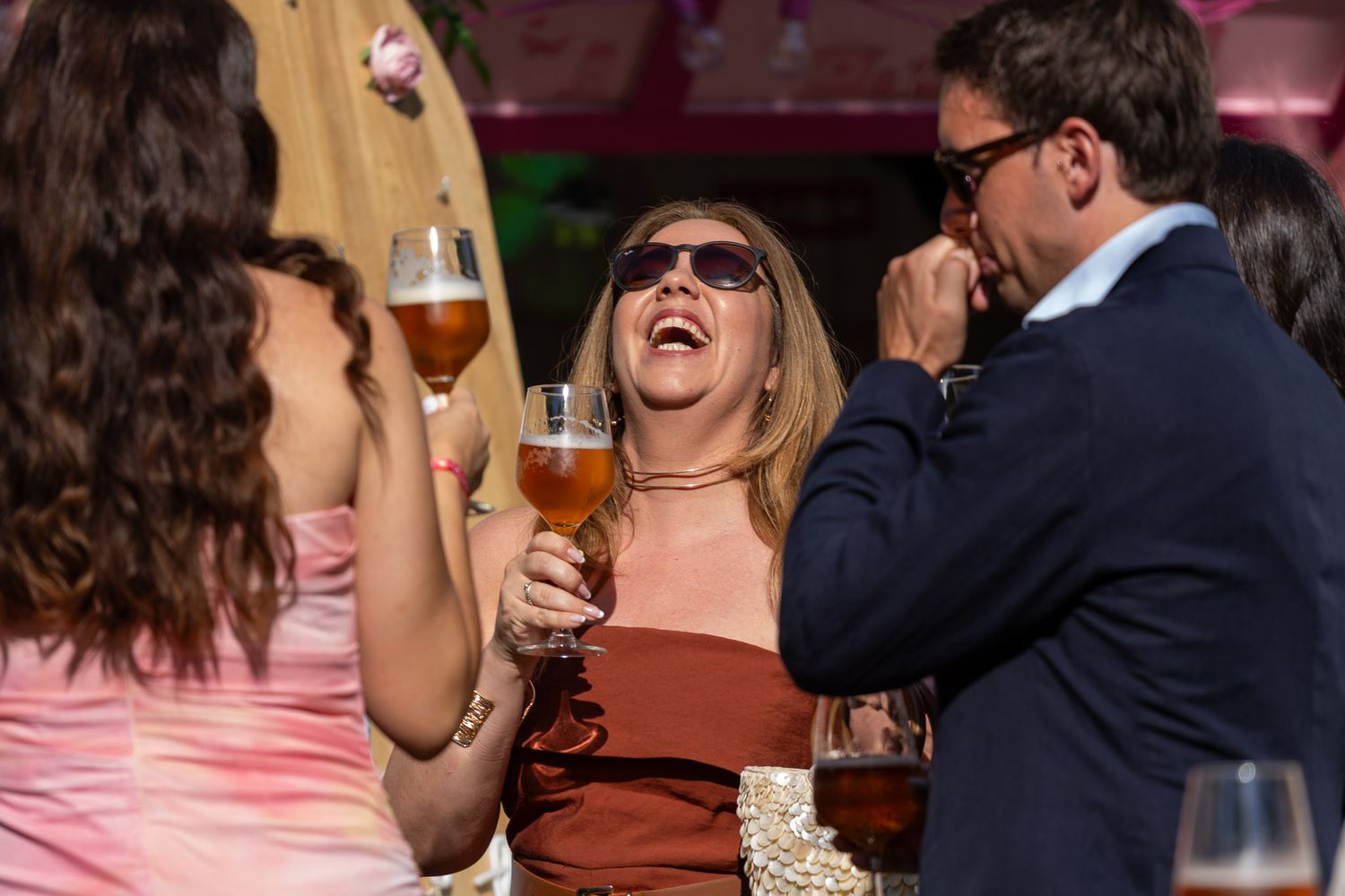 Three people laughing and socializing outdoors, holding glasses of beer at a sunny social gathering.