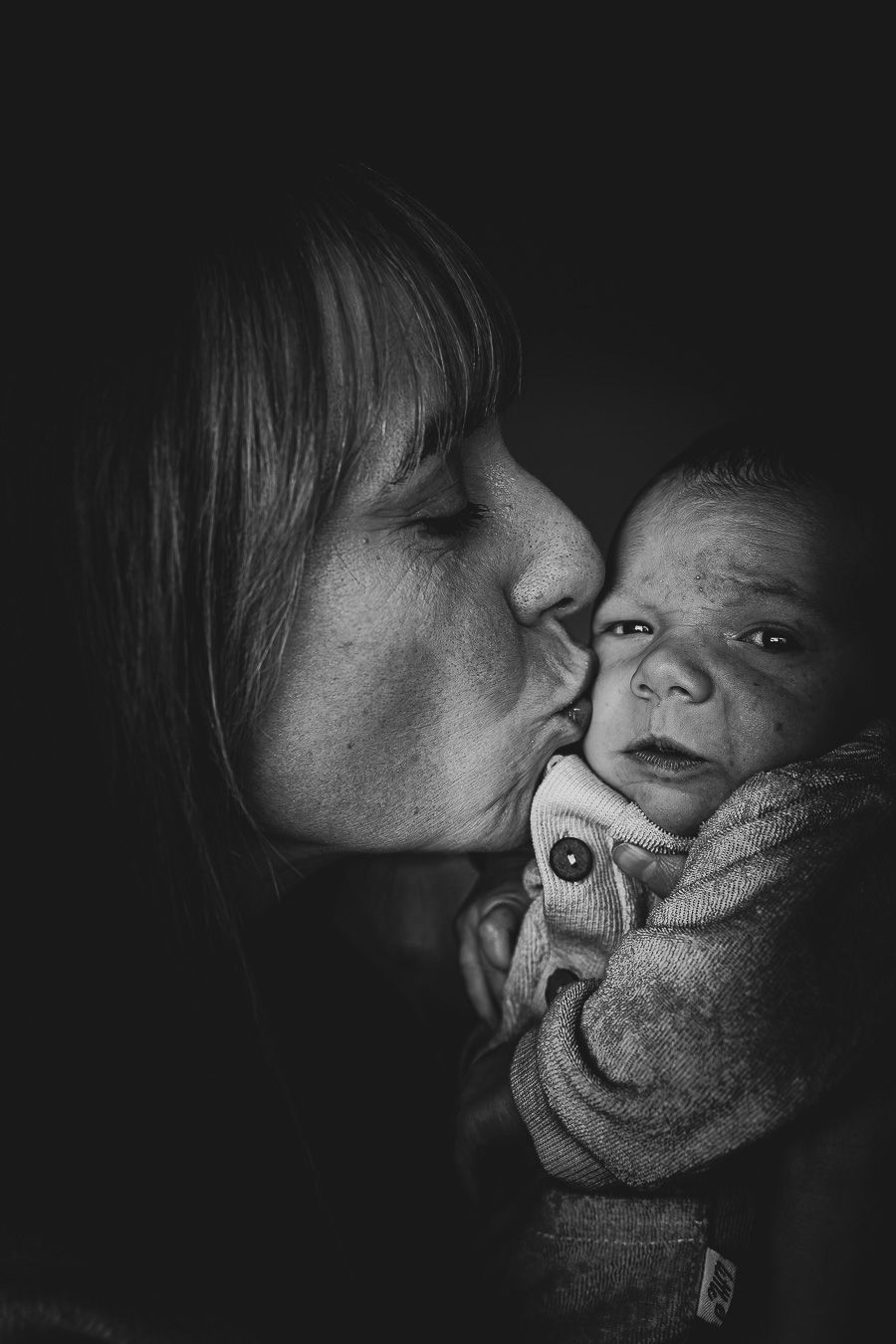 Black and white photo of a mother tenderly kissing her newborn baby, captured in a dark, intimate portrait.
