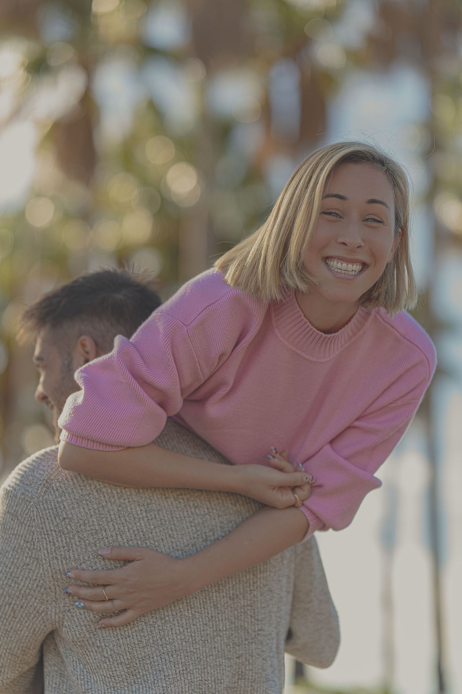 A smiling blonde woman in a pink sweater enjoys a piggyback ride outdoors near palm trees.