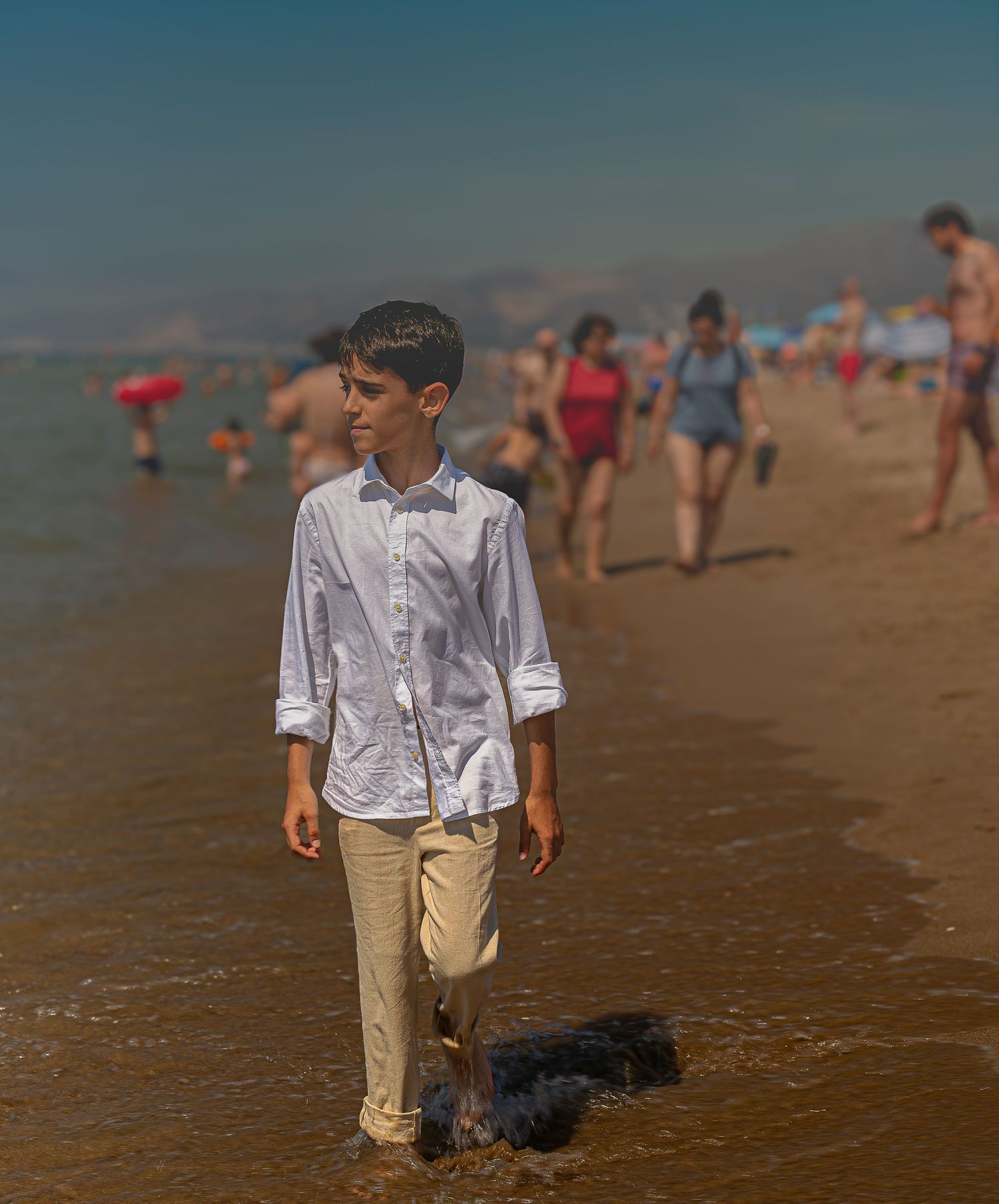 A young boy in a white shirt and khaki pants walks along a sunny beach shoreline with people in the background.