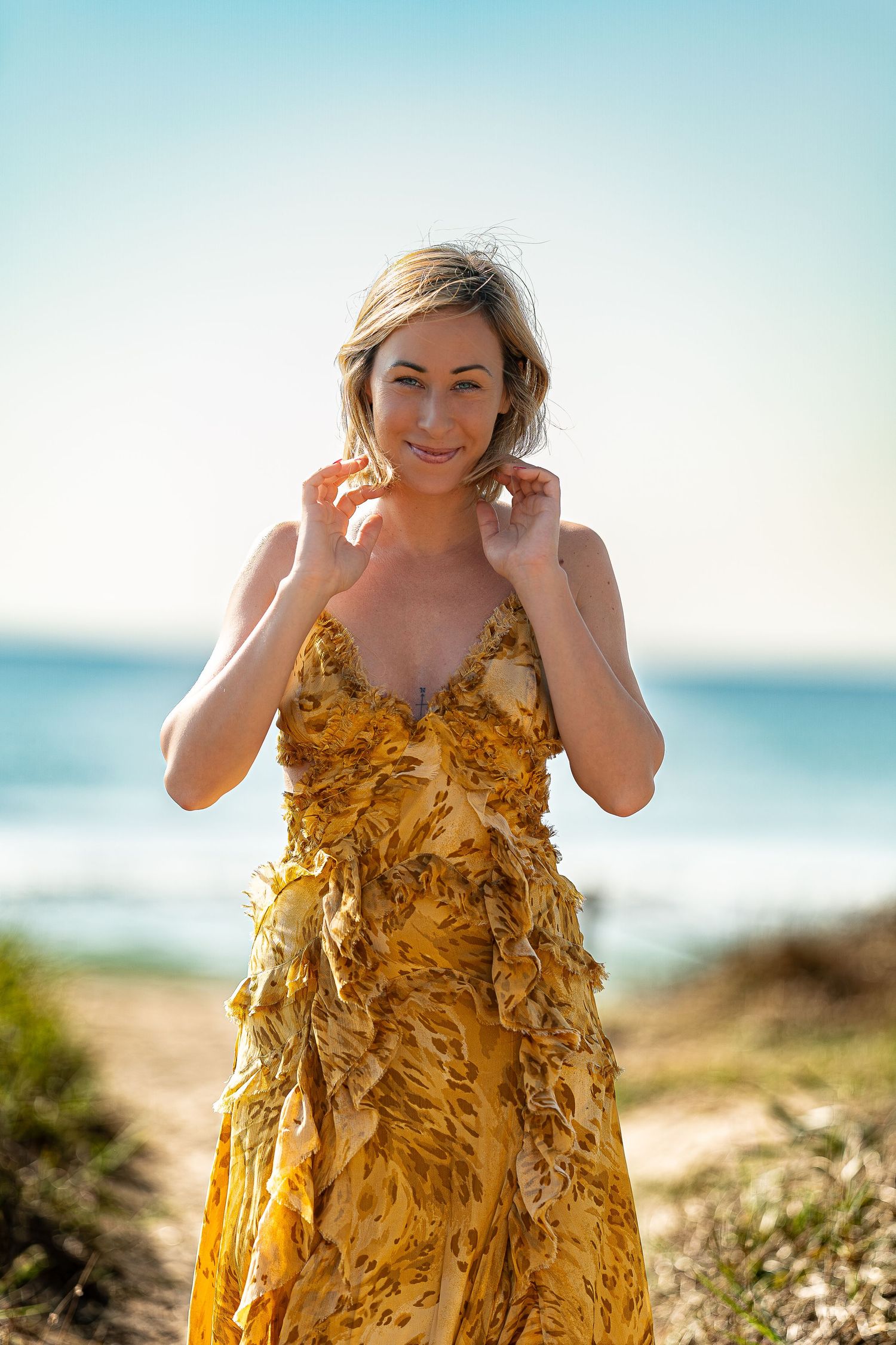 Una mujer con un vestido amarillo único hecho con hojas otoñales posa frente a un soleado paisaje costero.