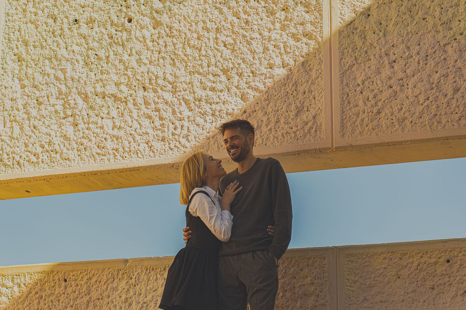 Pareja sonriendo y mirándose con complicidad frente a un muro urbano, sesión de fotos de pareja al aire libre en Barcelona, estilo Meraki.