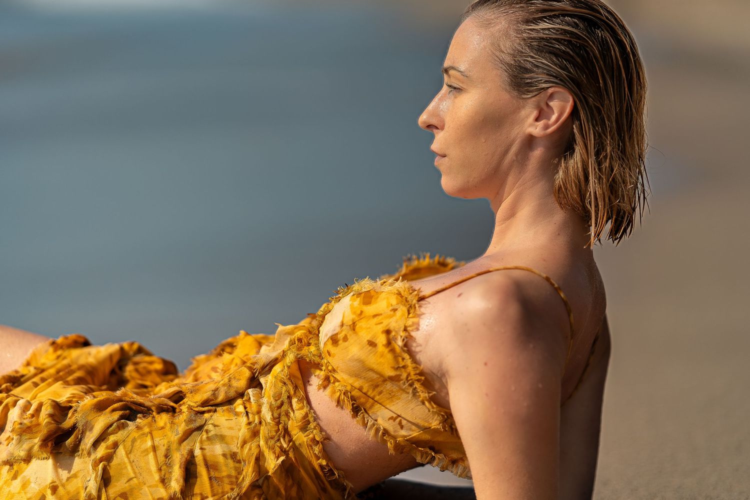 Woman in yellow ruffled dress sitting by the ocean, profile view with wet hair in golden sunlight.