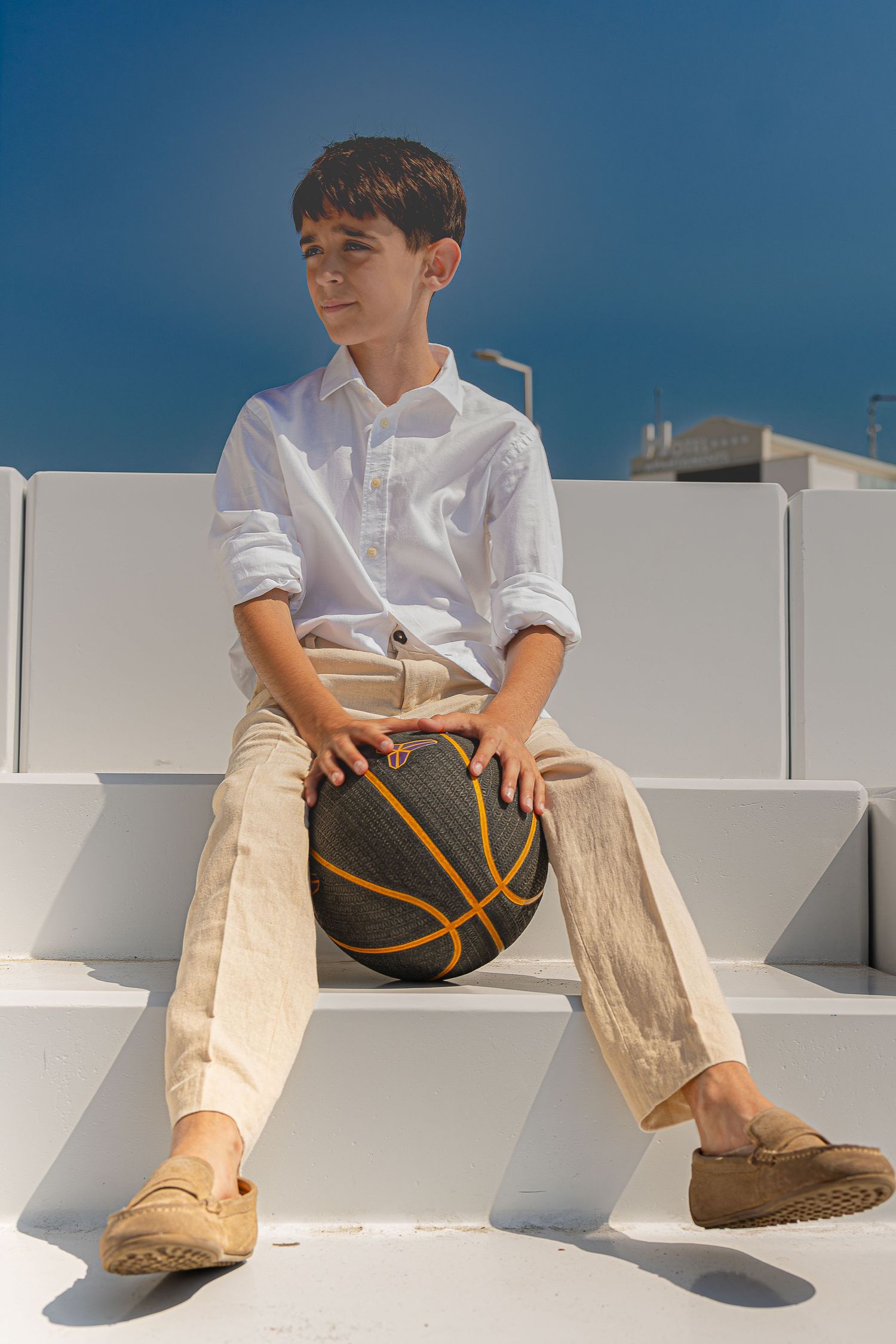Boy in white shirt and beige pants sitting on white steps holding a basketball on a sunny day.