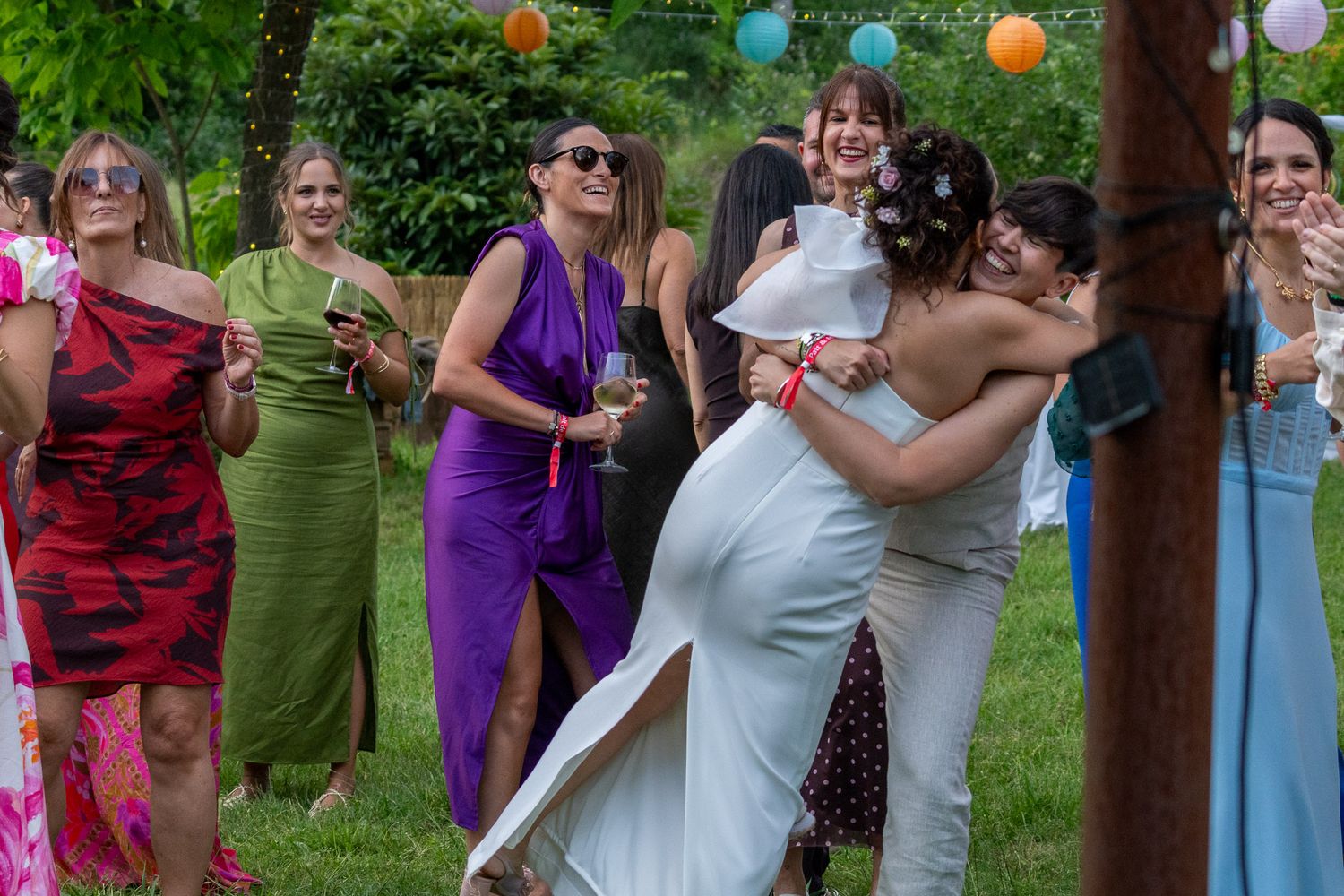 Bride hugging a guest at an outdoor wedding celebration with colorful dressed women laughing in background.