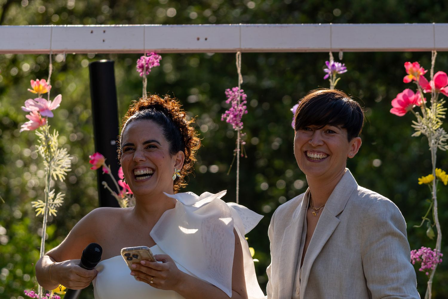 Two brides laughing joyfully under a floral arch with pink and yellow flowers at an outdoor wedding ceremony.